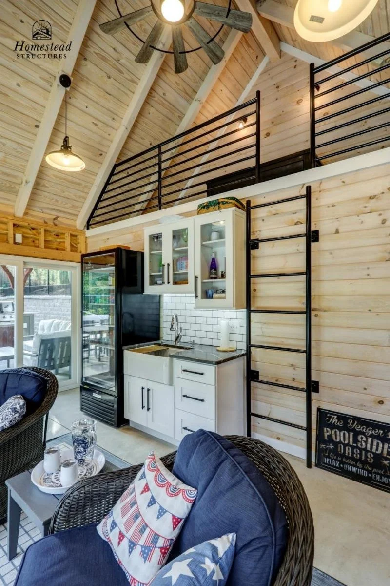 Interior view of a cozy, modern cabin with a high, wood-paneled ceiling, a black metal railing on a loft area, a small kitchen with white cabinets and a black refrigerator, wicker chairs with cushions, and a sliding glass door leading outside.