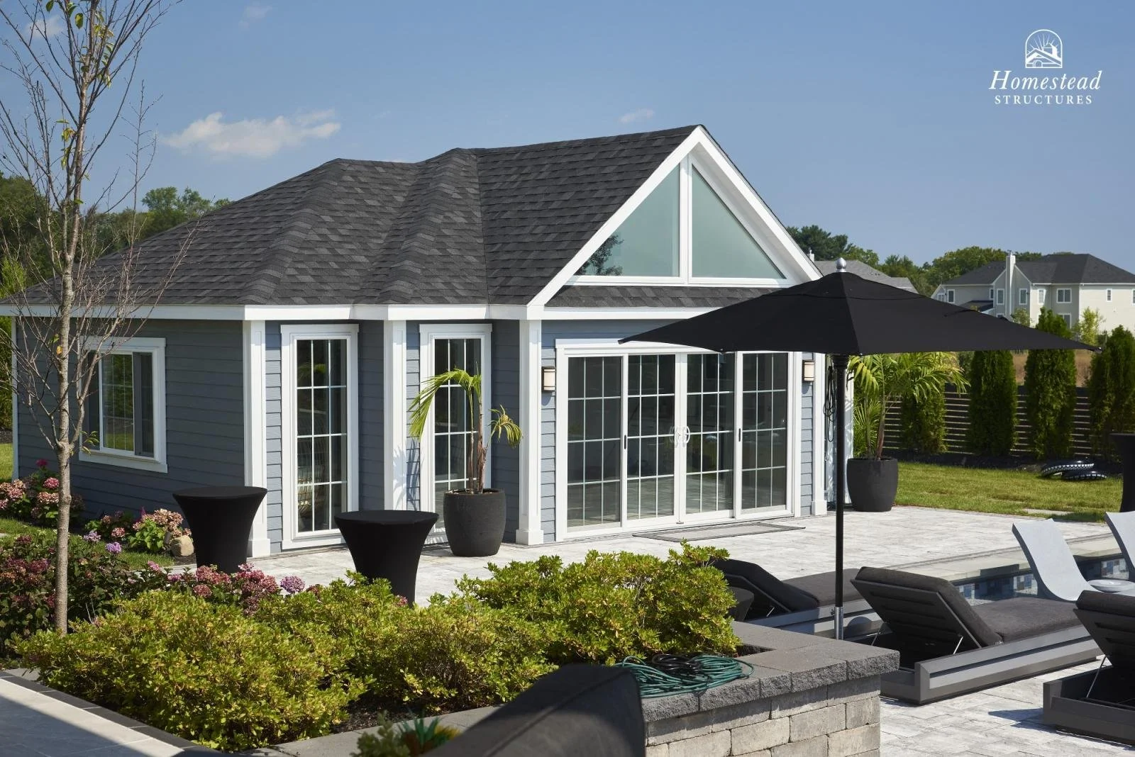 A modern backyard patio with lounge chairs, large black patio umbrella, potted plants, and a gray house with large windows and a triangular window on the roof, under a blue sky.