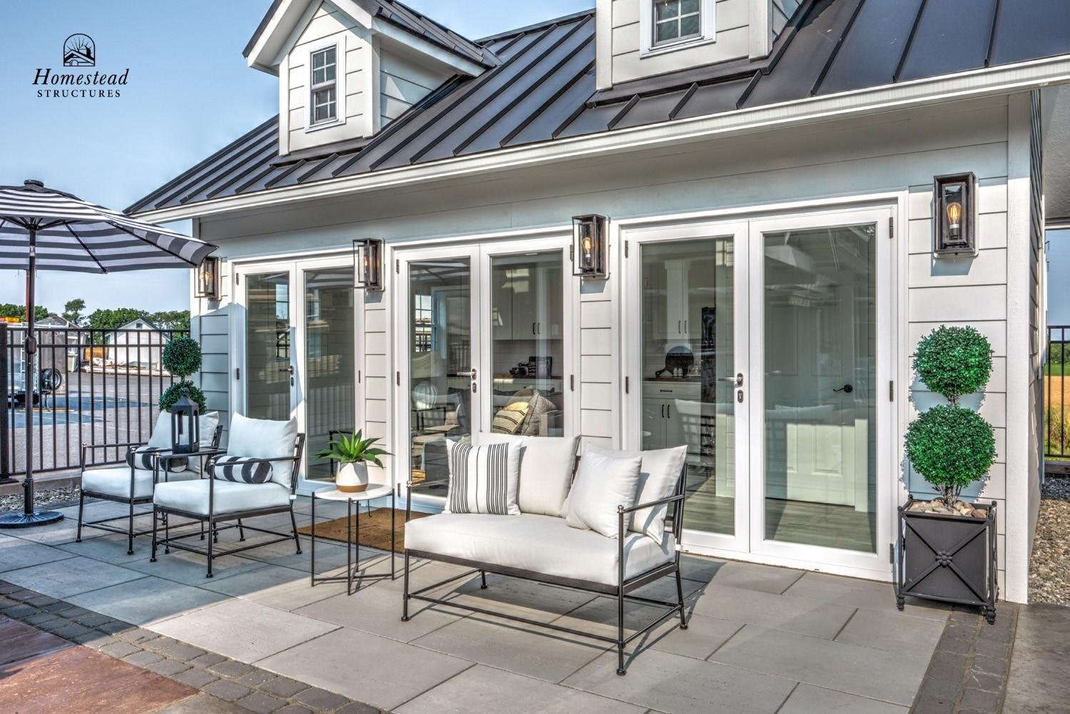 A backyard patio with white outdoor furniture, including a sofa, chairs with white cushions, and a black and white striped pillow. There are potted plants, a large umbrella, and a sliding glass door leading into the house. The house has white siding,