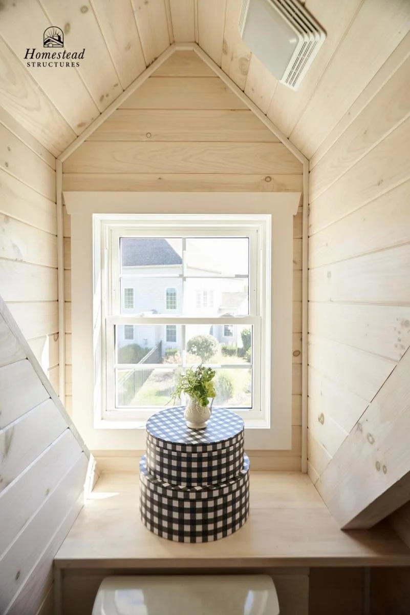 Small room with light wood paneled walls and ceiling, a window showing neighboring houses and yard, a table with a checkered black-and-white tablecloth, and a vase with green plants on top.