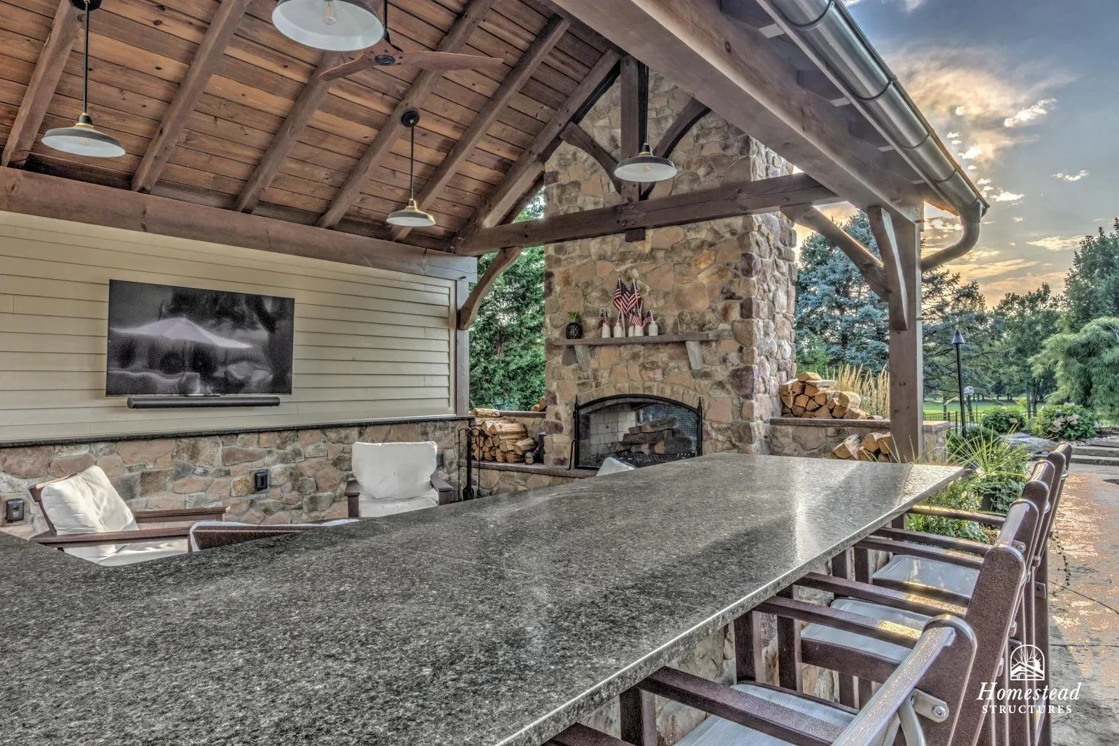 An outdoor patio with a stone fireplace, wooden ceiling, TV mounted on a beige wall, and stone walls. There is a long granite table with chairs and a view of trees.
