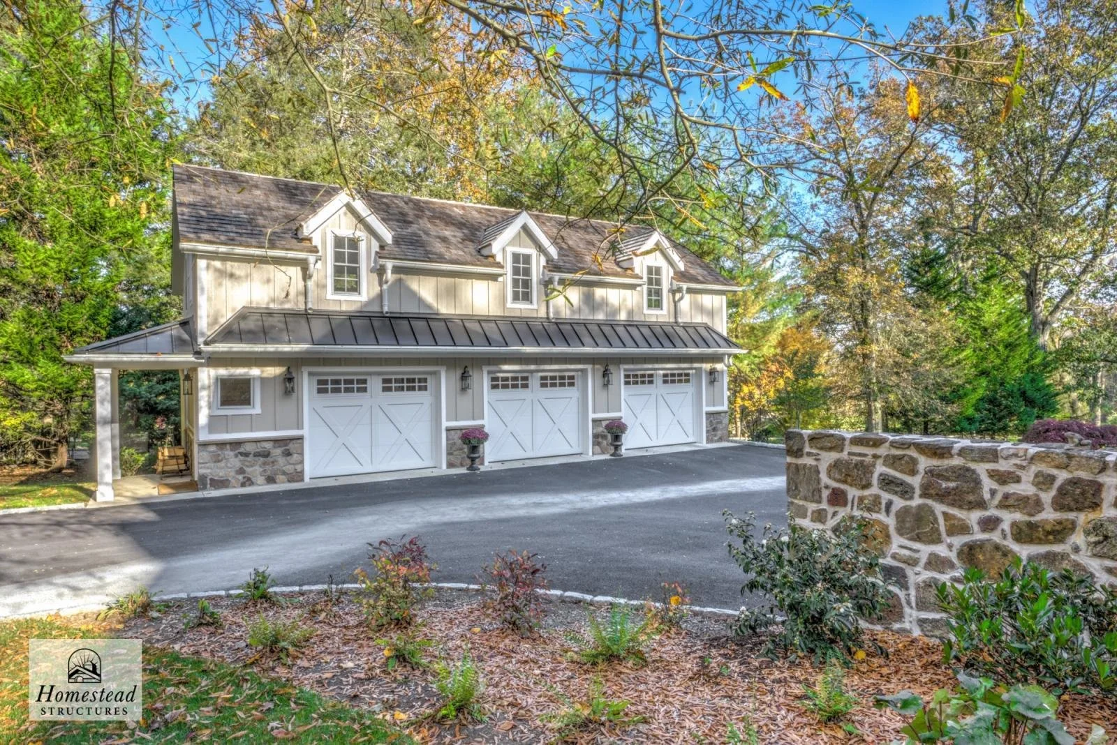 A detached three-car garage with a stone foundation and white garage doors, situated in a wooded area during daytime with fall foliage.