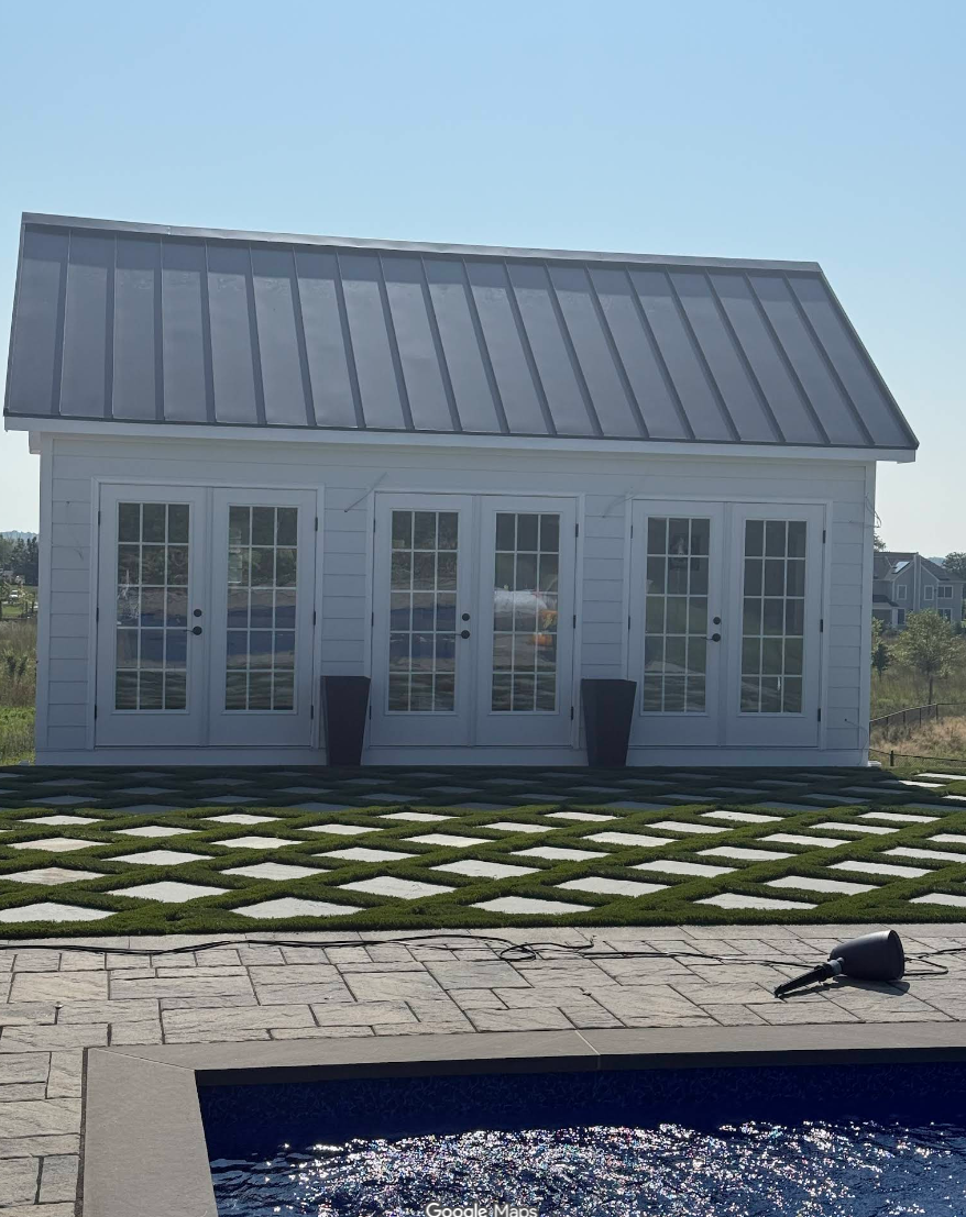 A small white building with four glass doors and a metal roof, set on a patterned lawn with squares of grass and concrete. A watering can lies on the patio near a pool.