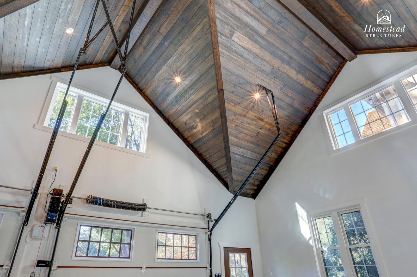 Interior of a spacious room with a vaulted dark wood ceiling, multiple large windows allowing natural light, and exposed garage door mechanisms.