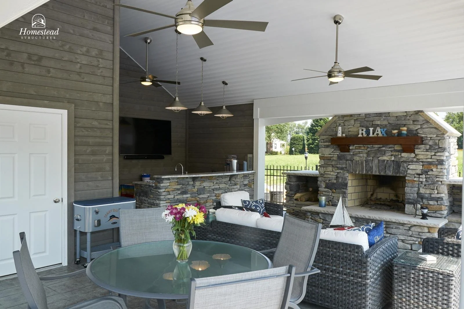 Covered outdoor patio with a stone fireplace, a round glass dining table with four chairs and a flower vase, wicker seating with cushions, a TV mounted on the wall, ceiling fans, and a bar area with lighting fixtures.