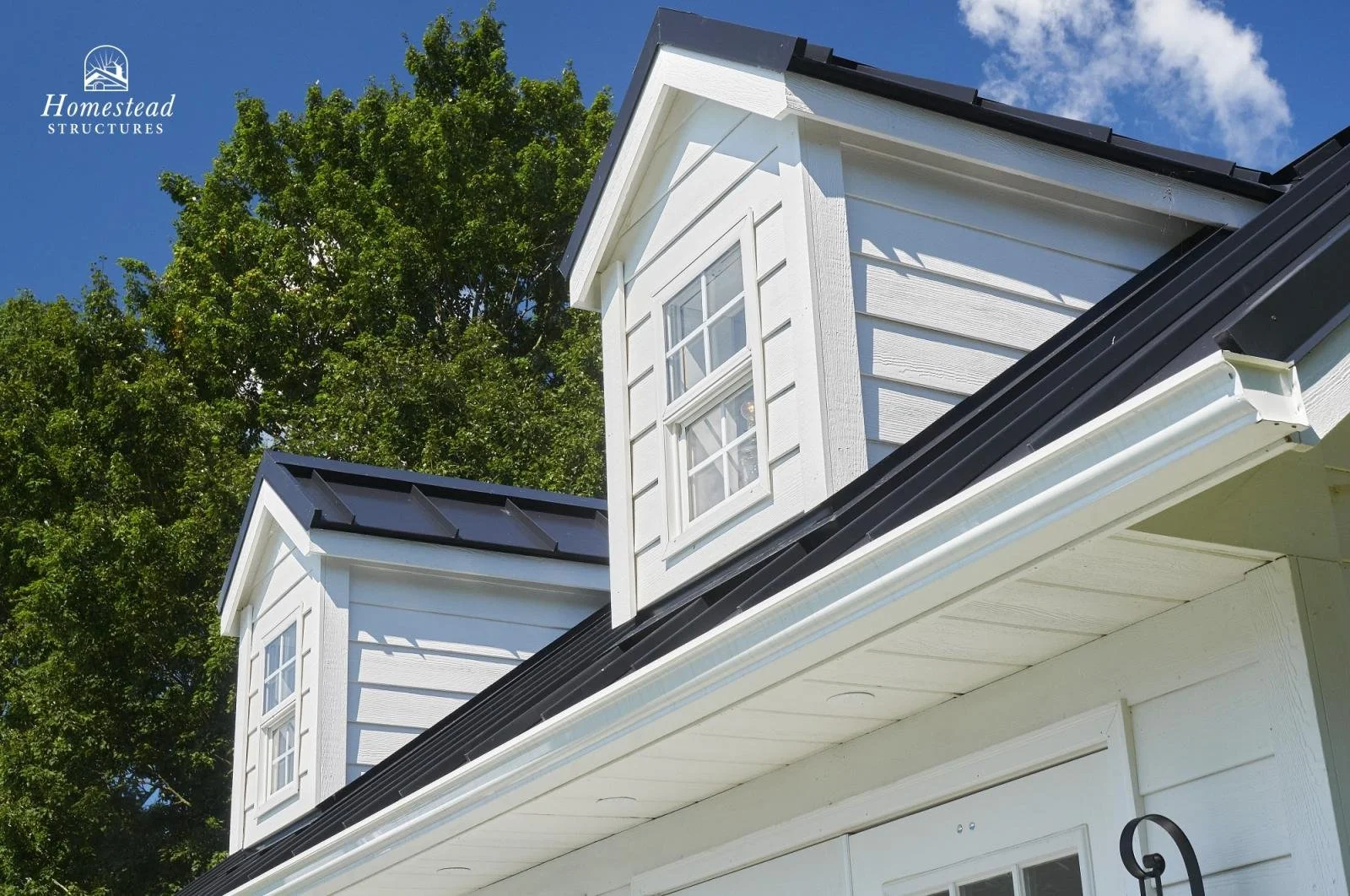 Close-up of a white house with two dormer windows, black roofing, and trees in the background.