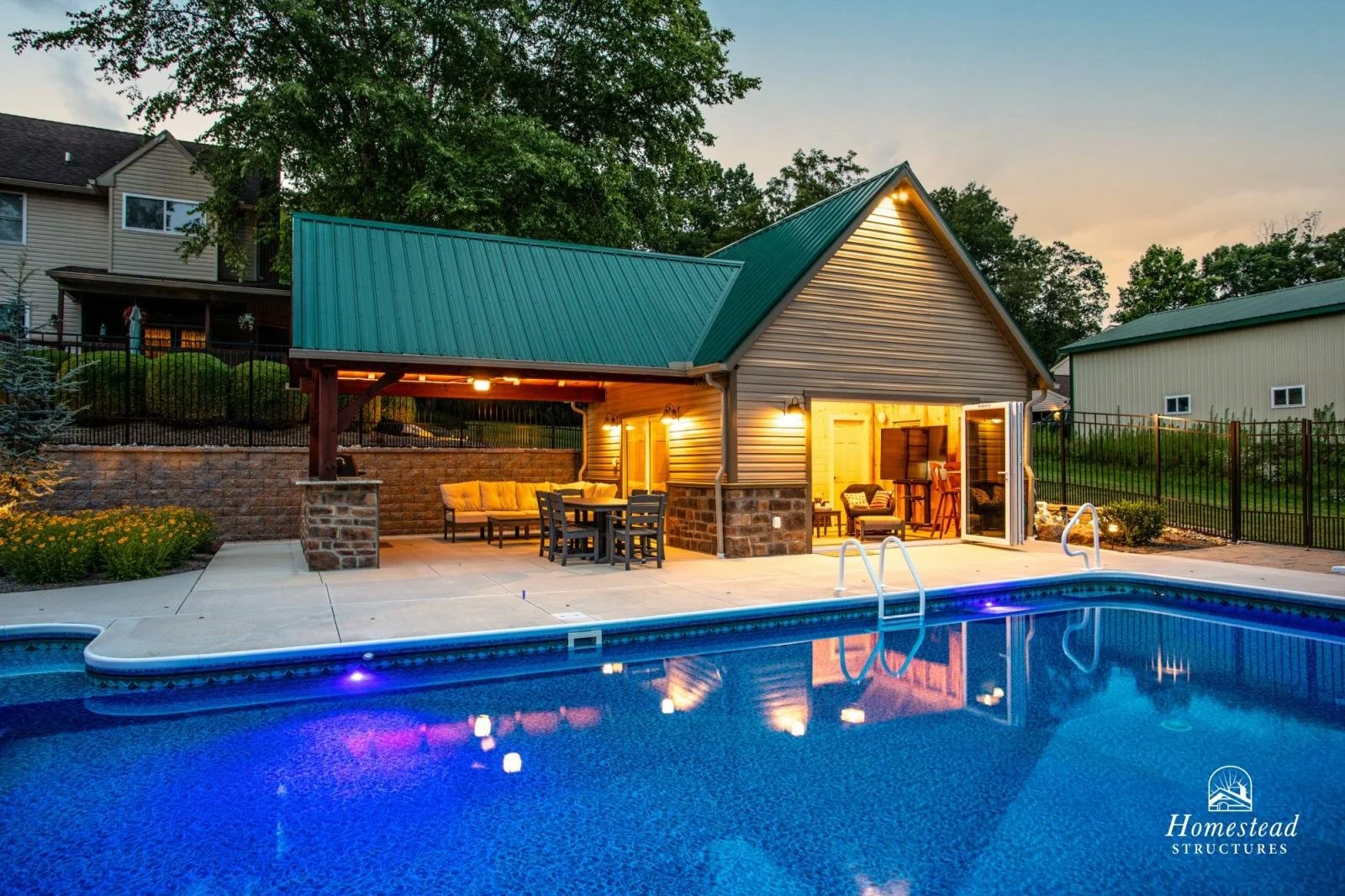 Backyard with a swimming pool, patio furniture, a small building with a green metal roof and outdoor lighting at dusk, surrounded by a fence and trees.