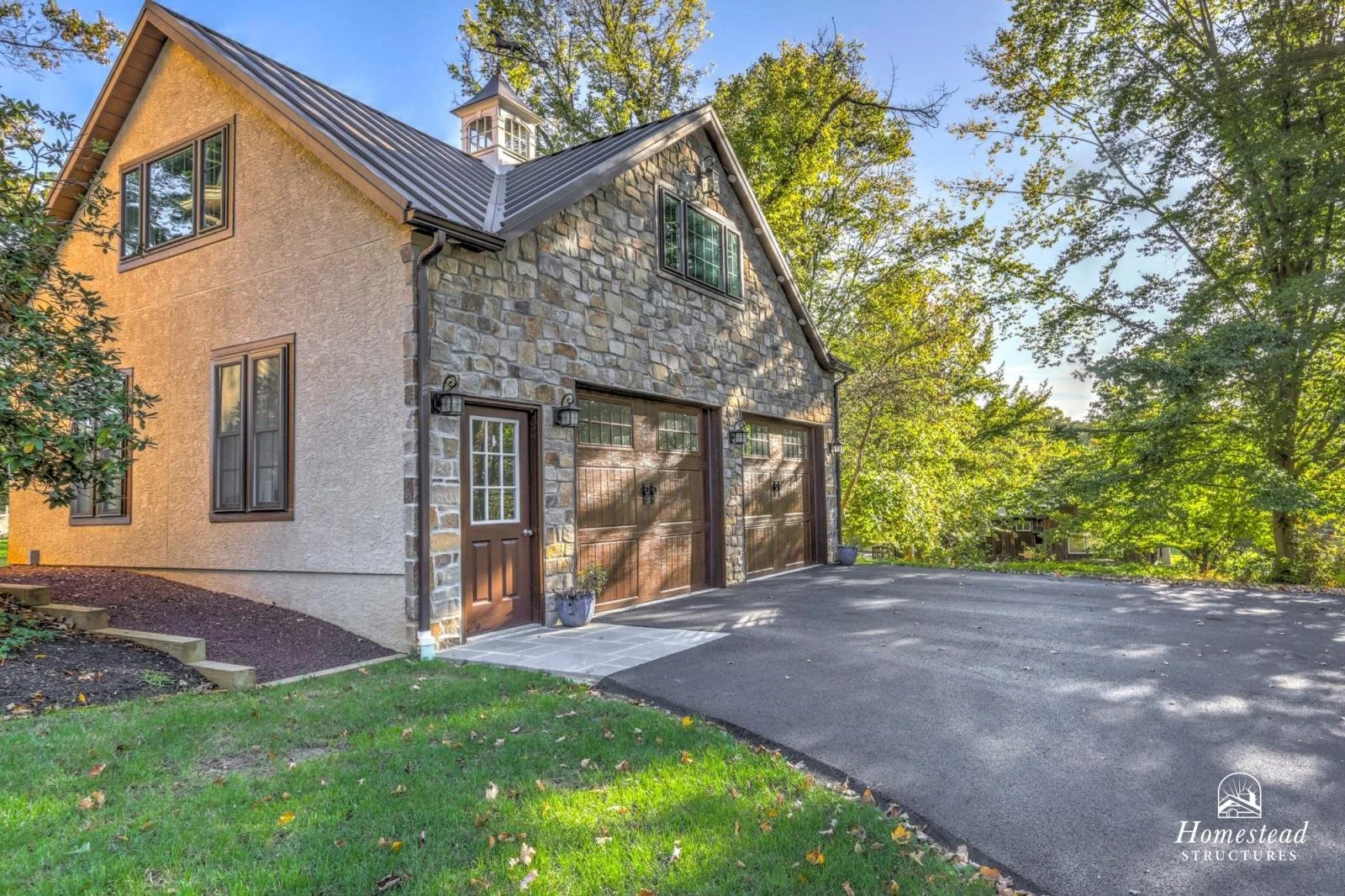Exterior of a house with a stone and stucco facade, brown garage doors, and a paved driveway surrounded by green trees and grass.