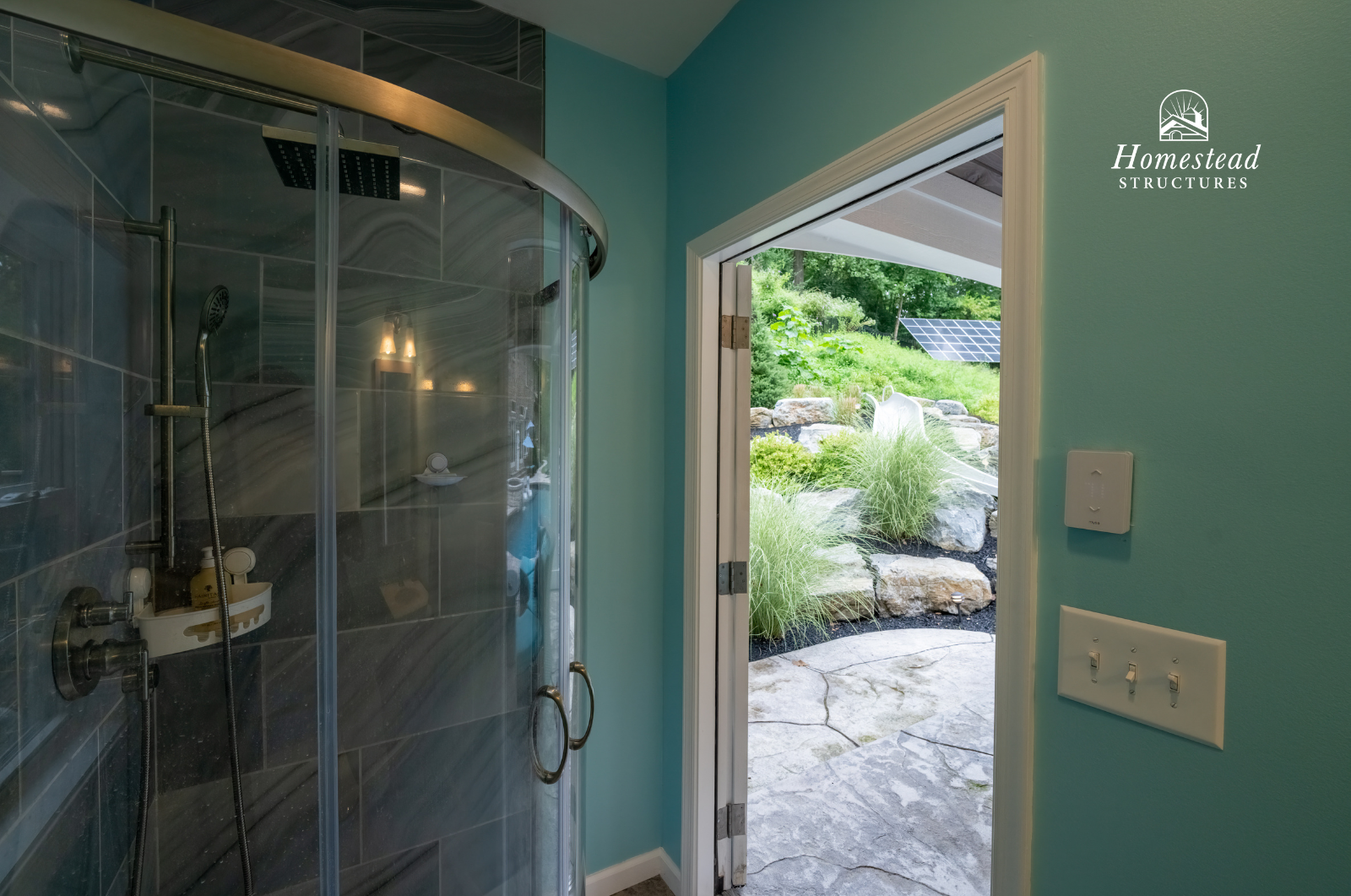 Indoor shower with glass door open to an outdoor garden area with rocks and greenery.