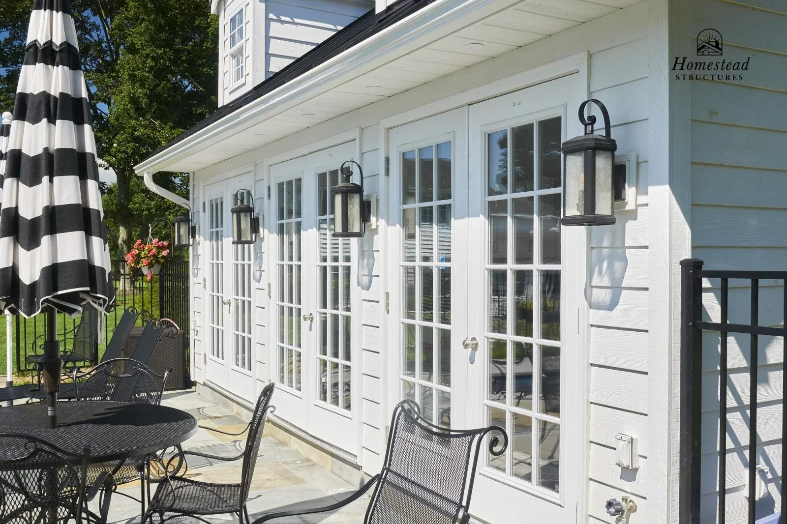 Exterior of a house with French doors, black metal outdoor furniture, a striped black and white umbrella, and lantern-style wall lights