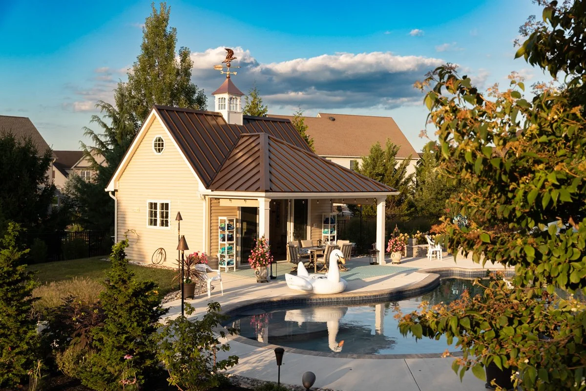 Backyard with a small house, swimming pool with a swan-shaped float, outdoor furniture, trees, and a clear blue sky.