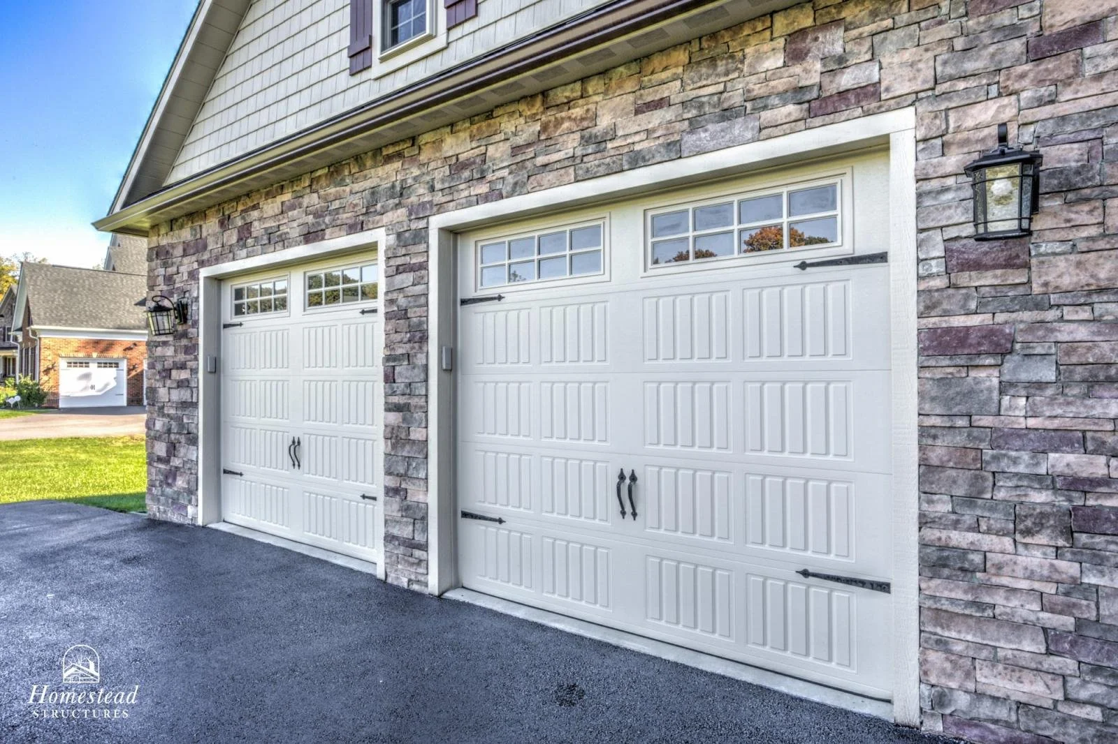 Two white garage doors with small windows at the top, attached to a house with stone and siding exterior, with black lantern-style lights on each side.