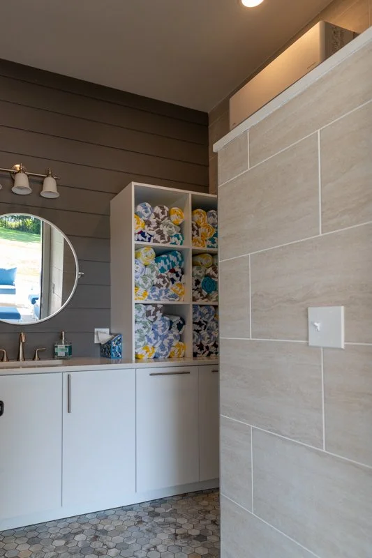 Bathroom with cabinet filled with colorful rolled towels, round mirror, and bathroom sink with soap and toiletries, beige tiled wall, and hexagonal tiled floor.