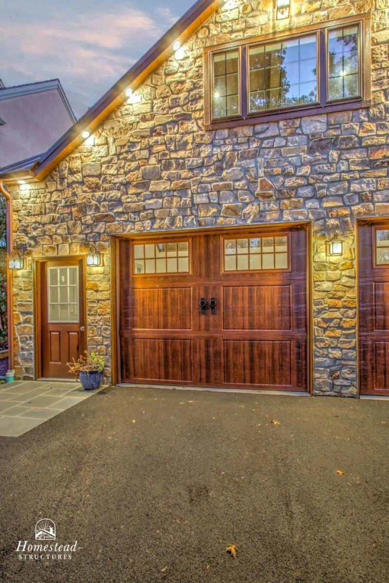 A stone house with a wooden garage door and side door, exterior lights, and a small plant in a pot by the side door, taken during the evening.