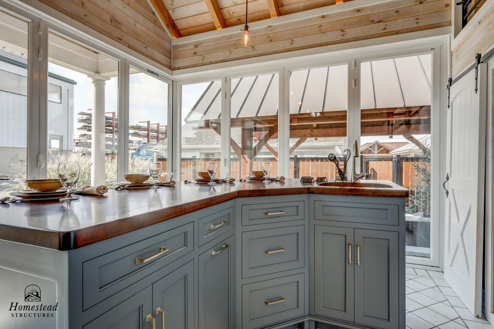 Interior view of a kitchen with large windows, gray cabinets with gold handles, a wooden countertop, and a small sink. The ceiling is wood-paneled with a hanging light fixture. Outside the windows, there is a patio with a wooden pergola and neighbori