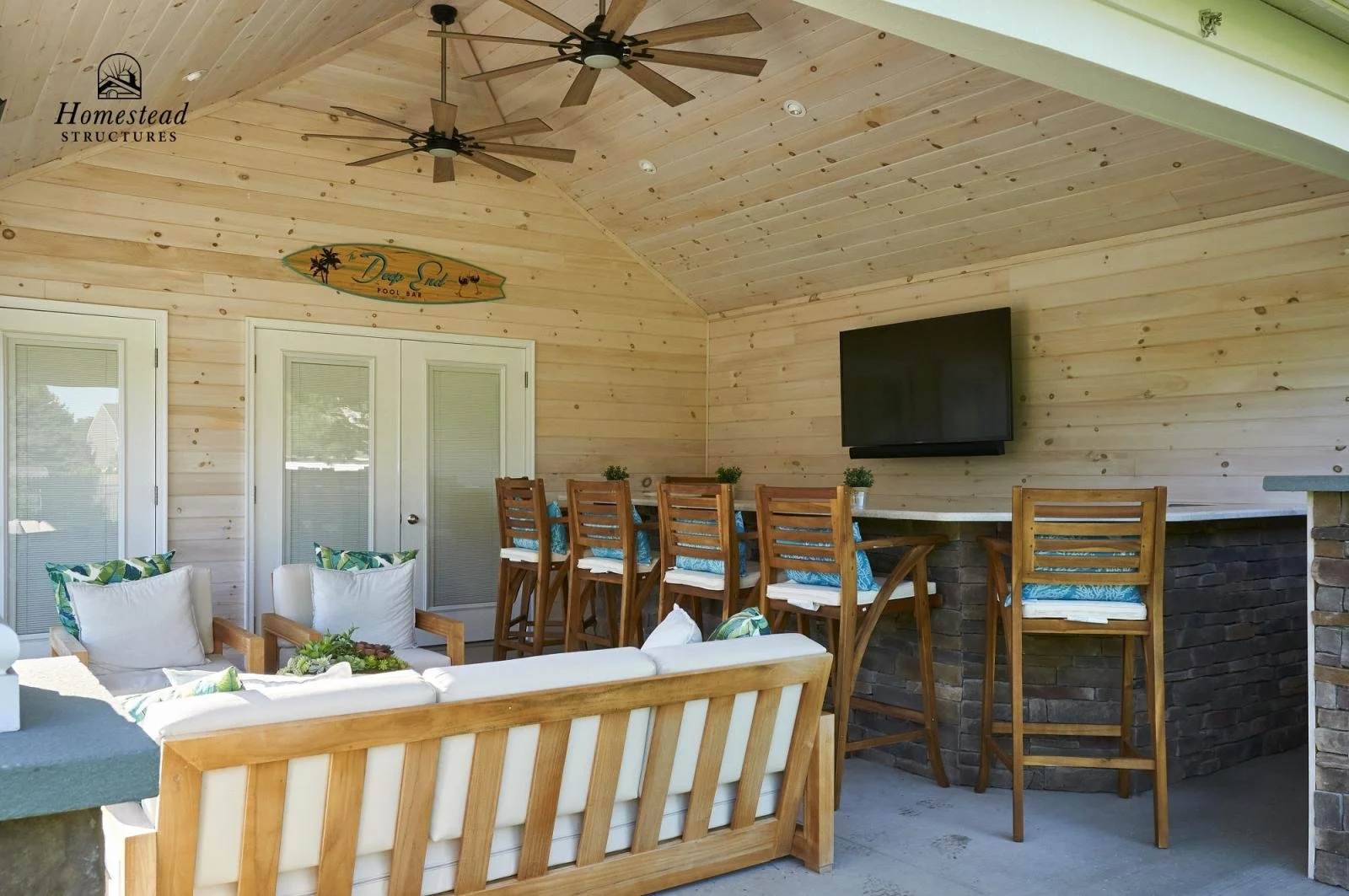 Outdoor patio with wooden furniture, white cushioned sofas, bar counter with wooden chairs, a mounted TV, and a wood-paneled ceiling with ceiling fans, in a backyard setting.