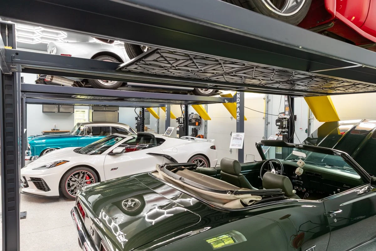 Interior view of a car storage facility with multiple classic and sports cars, including a green convertible in the foreground, a white sports car, and a vintage teal car.