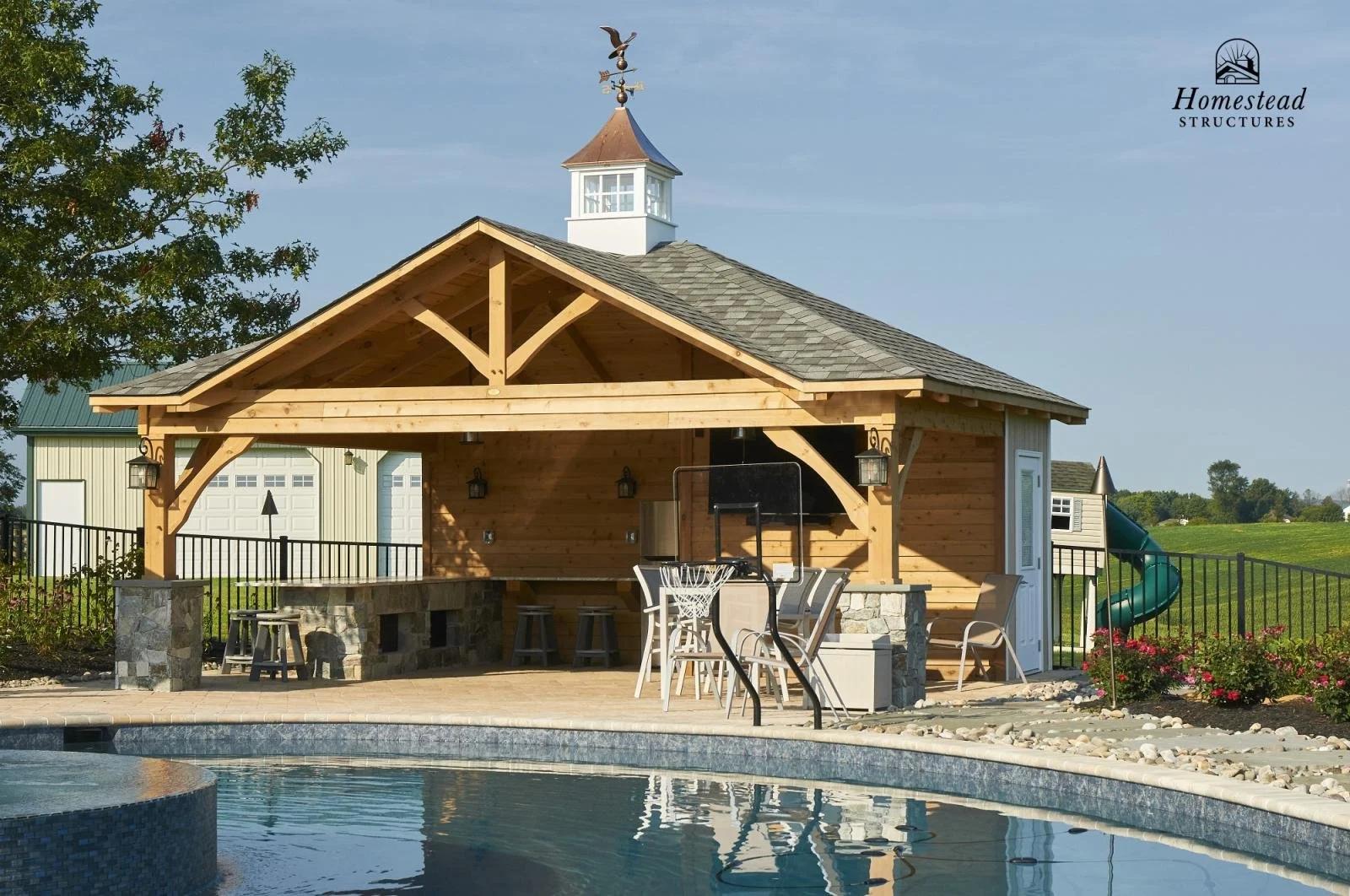 Wooden poolside cabana with a shingled roof, outdoor seating, and a basketball hoop next to a swimming pool, with a waterslide in the background and landscaping including flowers and trees.