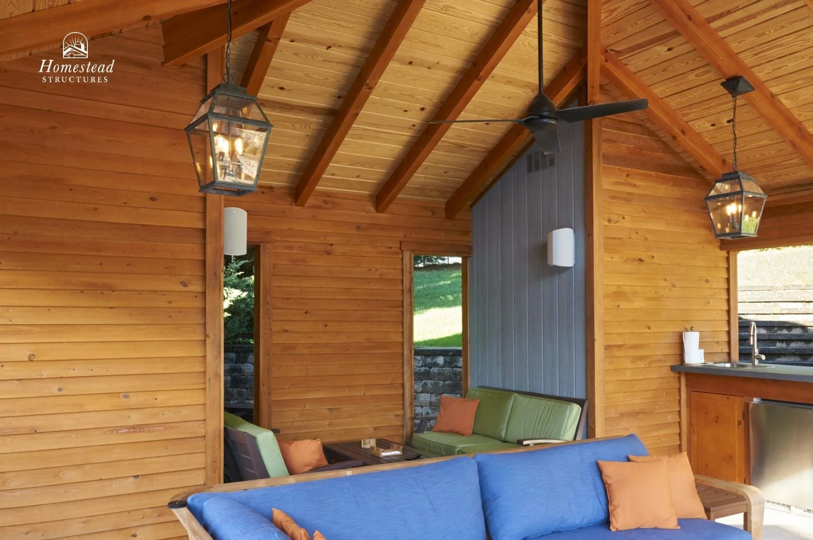 Interior of a wooden outdoor living space with a sloped wood ceiling, two lantern-style pendant lights, a ceiling fan, and a blue sofa with orange pillows. A partial view of a kitchen counter with a faucet is visible.