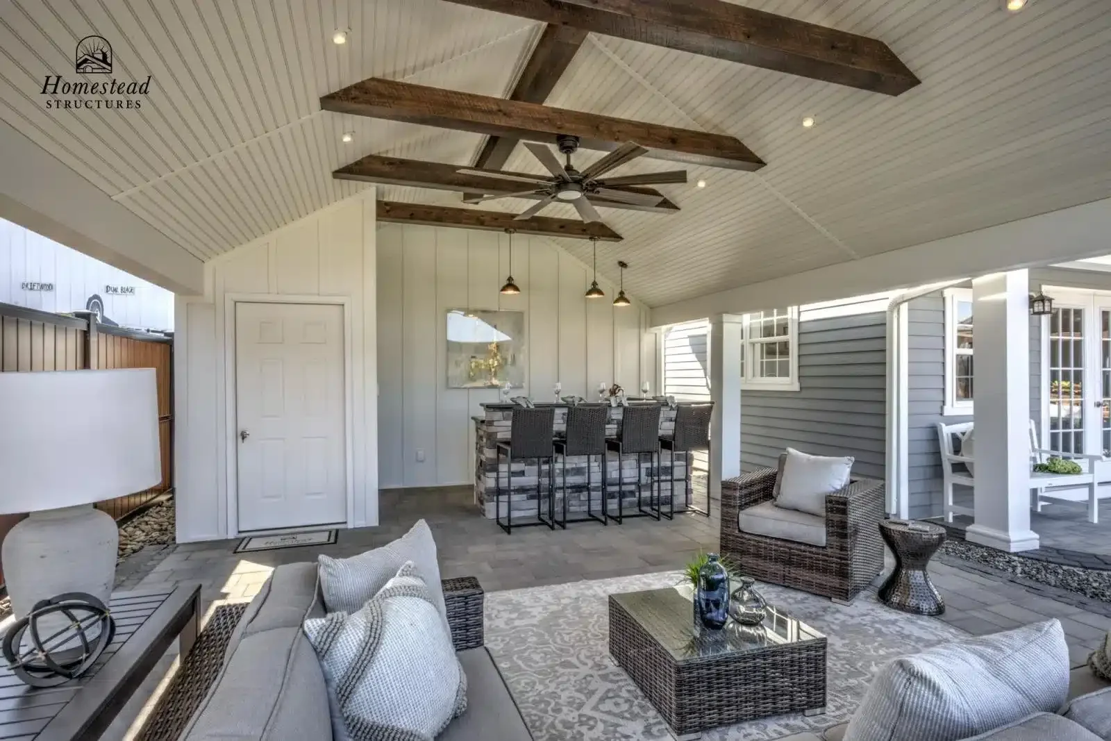 Covered outdoor patio with seating area, bar, and dining space featuring wicker furniture, a rug, a stone bar, and decorative accessories under a ceiling with exposed wooden beams and a ceiling fan.