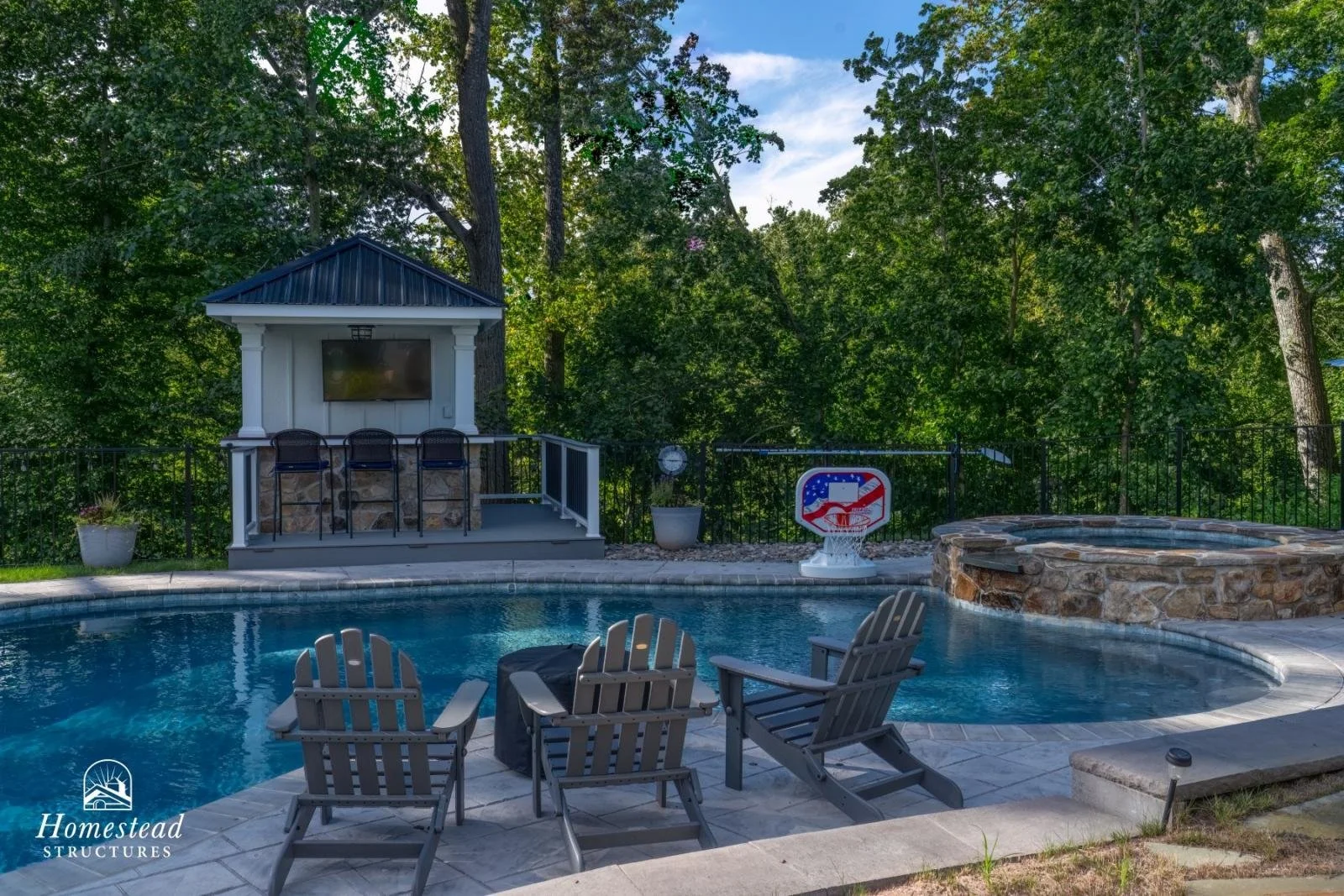 Backyard with a swimming pool, hot tub, lawn chairs, a pond, and a small building with a TV and three chairs, surrounded by trees.