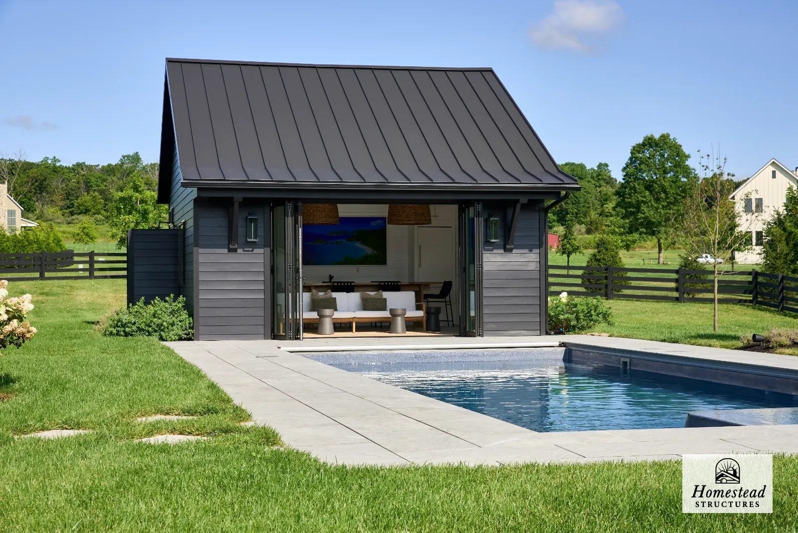A modern backyard with a dark gray pool house featuring a steep metal roof and sliding glass doors open to a seating area with a white sofa and green pillows, surrounded by a lush green lawn and a rectangular swimming pool.