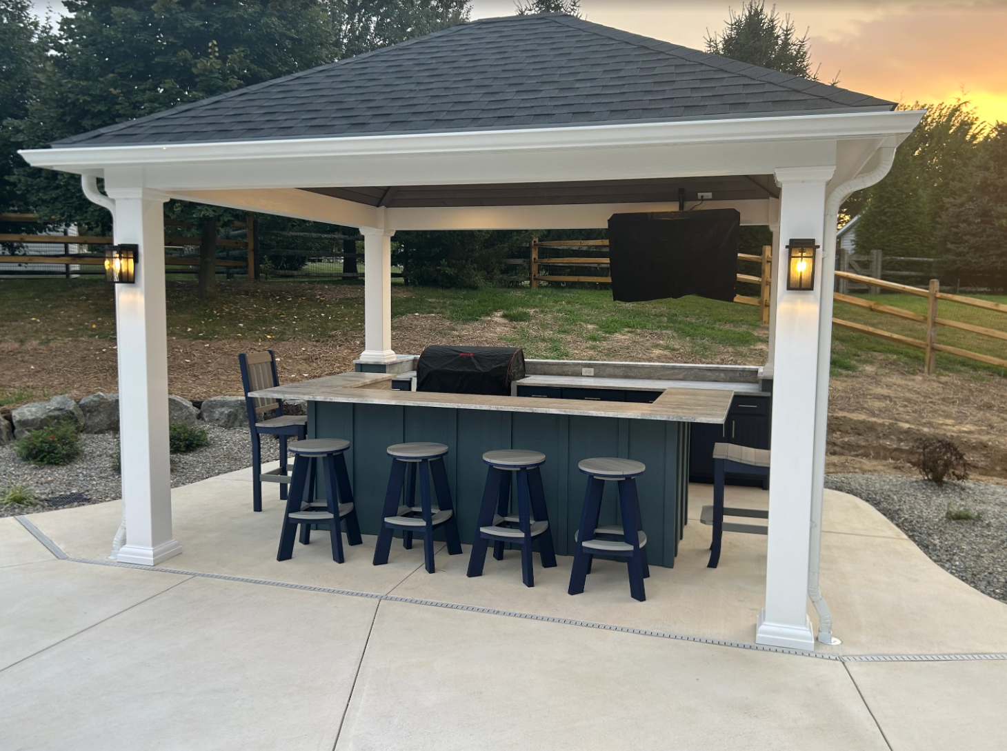An outdoor bar area under a pavilion with a dark roof, white pillars, and wall-mounted lights, with a counter, blue stools, a grill, and a television in the background, during sunset.