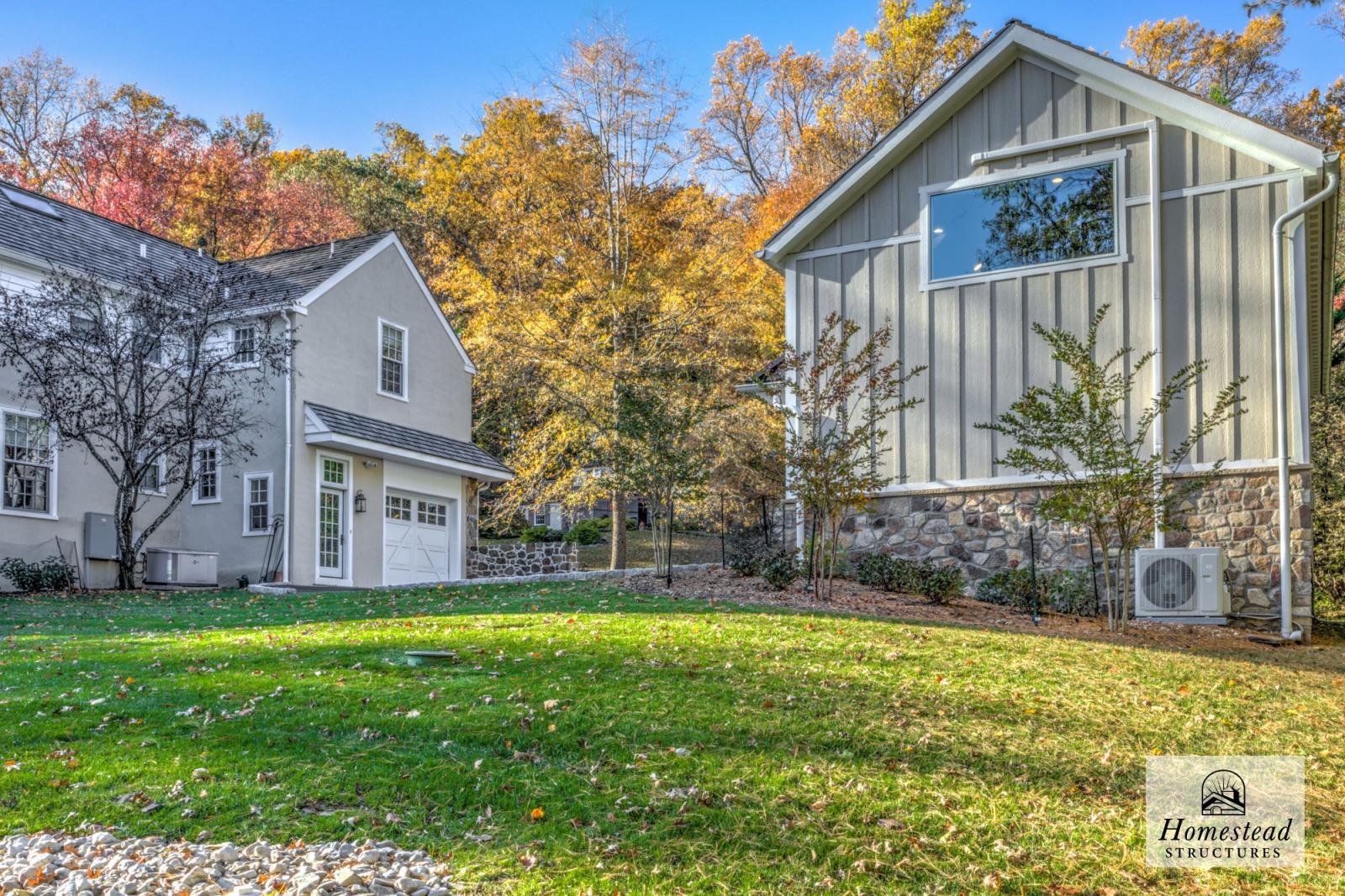 View of a backyard with a grassy lawn, several small trees, and two houses with different siding and roofing on a fall day with colorful foliage