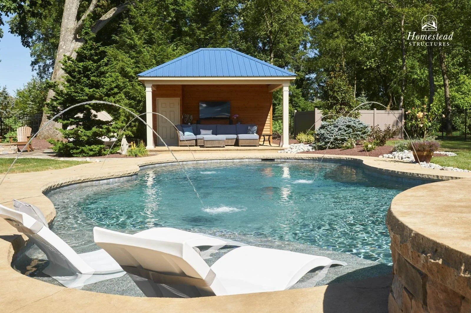 Backyard swimming pool with water features, lounge chairs, and a shaded outdoor seating area with a television, surrounded by trees and landscaped garden.