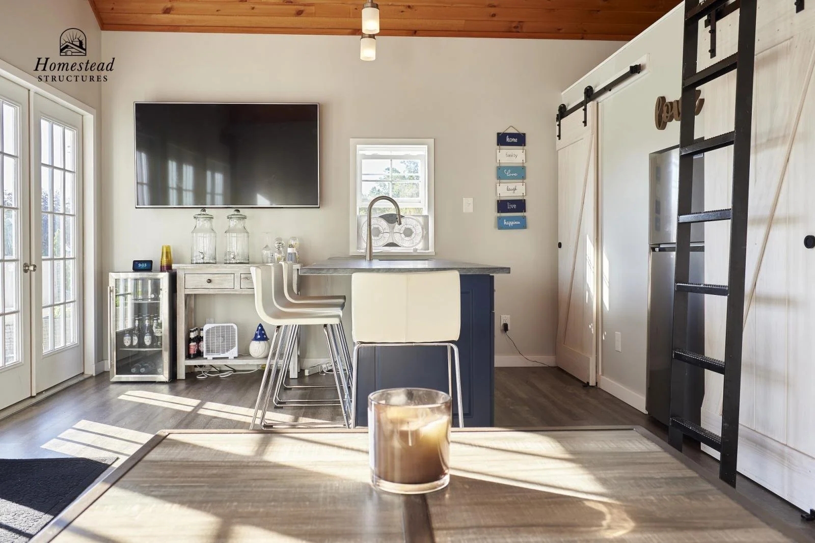 Kitchen with wooden ceiling, white walls, and sliding barn doors. Features a large black TV, small white table with bar stools, candles, a small fridge, a window with a fan, and wall decor. In the foreground, a candle on a wooden table.