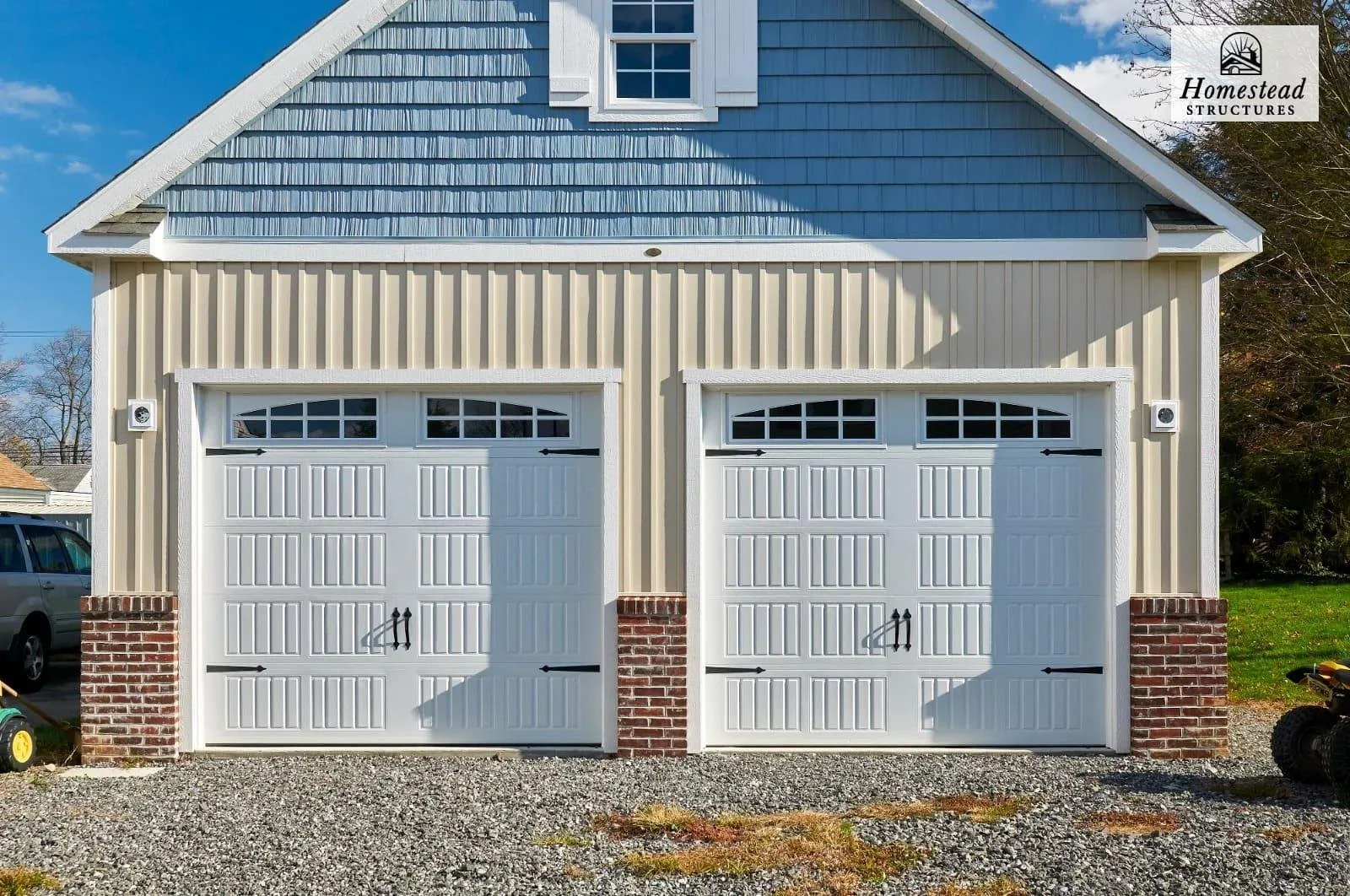 A two-car garage with white paneled doors featuring small windows at the top, brick accents at the bottom, in a building with beige siding and blue shingles on the upper part. There is a gravel driveway front and some objects on the sides, with a par