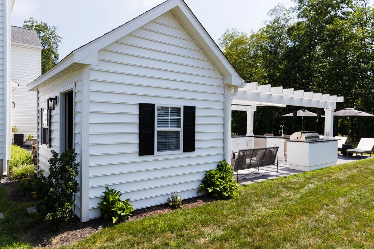 Backyard with a white house, outdoor kitchen, and seating area under a pergola, surrounded by green trees.