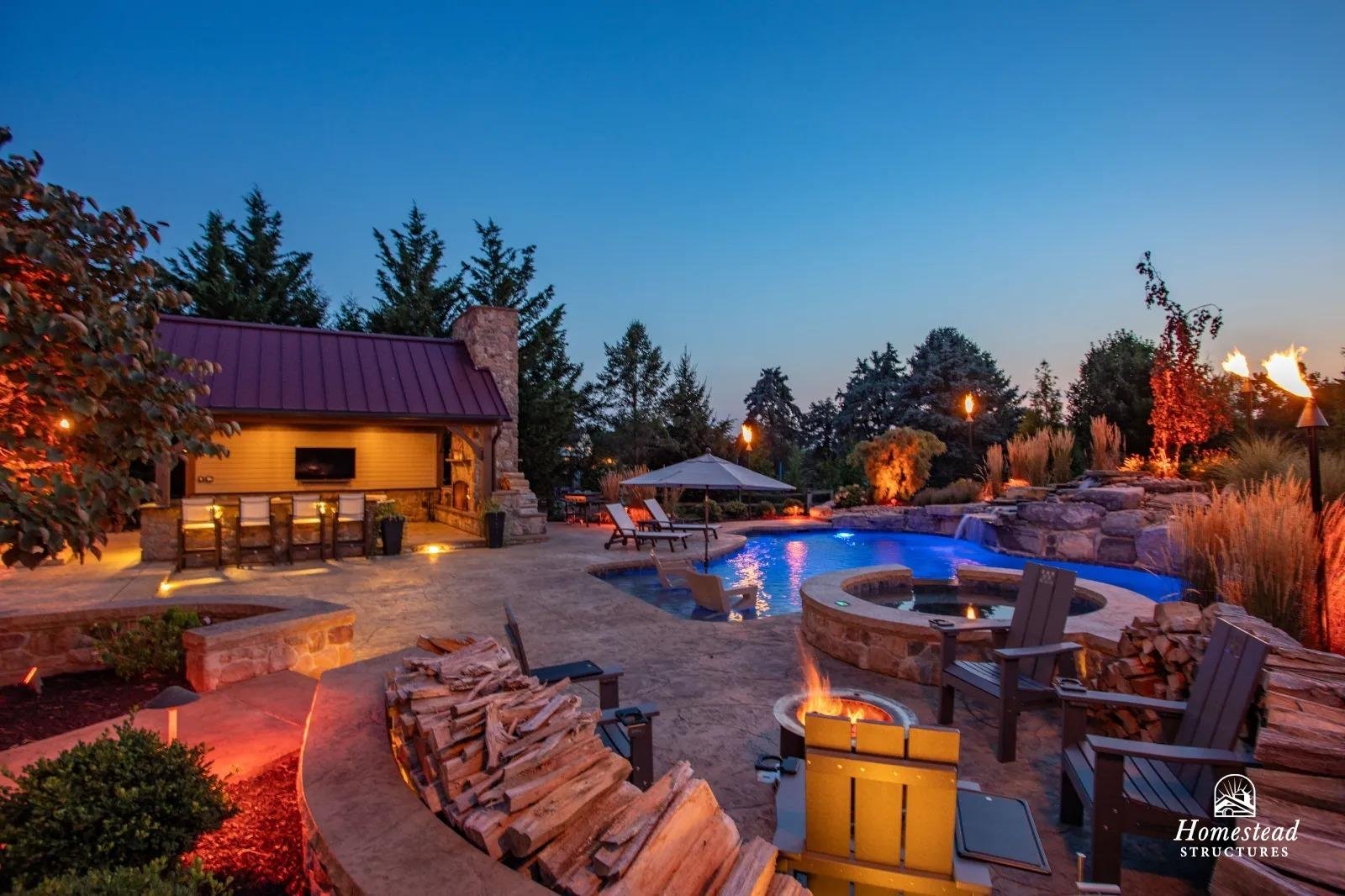 A backyard with a swimming pool surrounded by landscaping, fire pits, chairs, a covered patio, and trees, lit with outdoor lighting during dusk.
