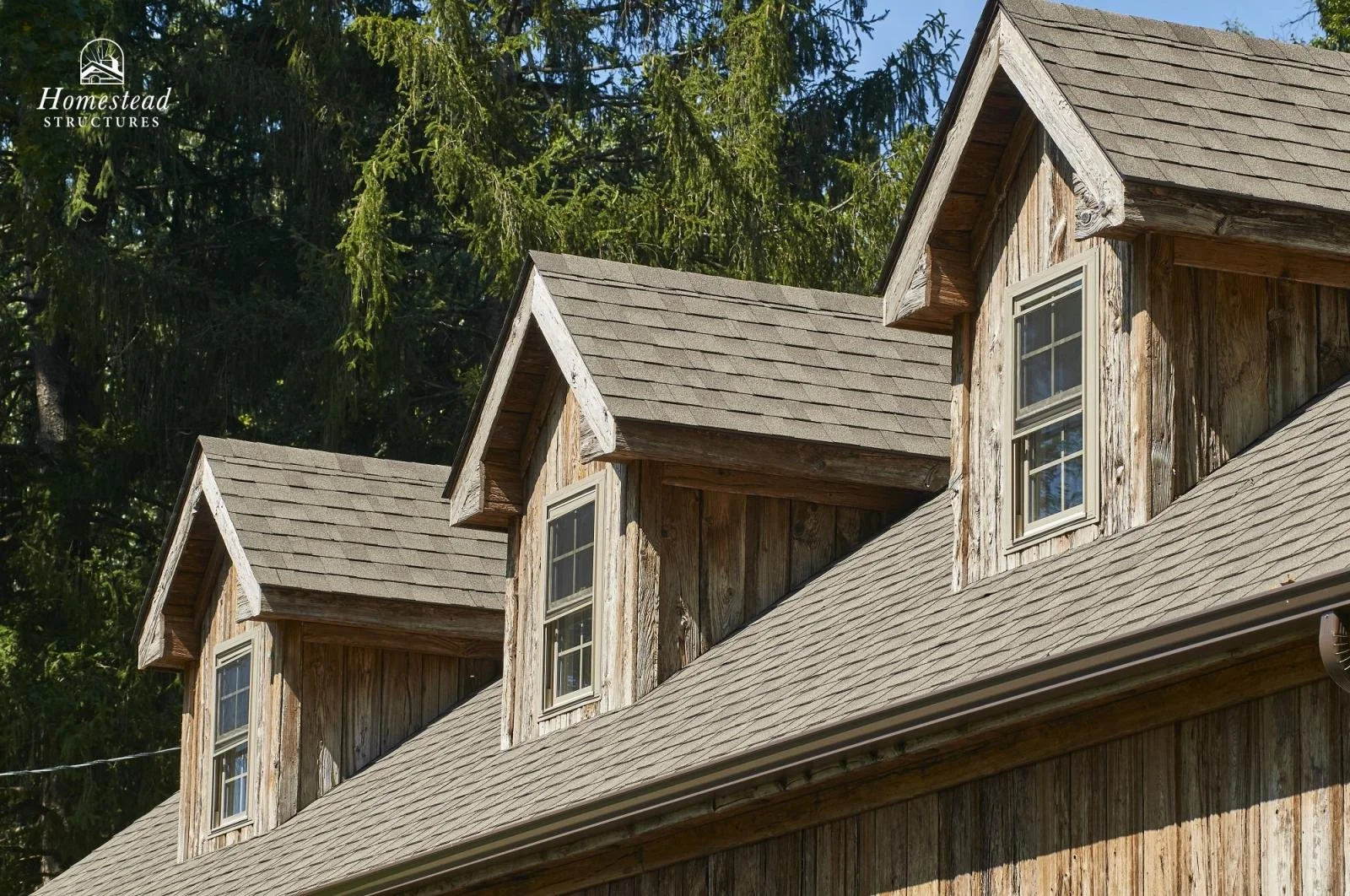 Close-up of a wooden barn with three dormer windows on the roof, surrounded by tall trees in the background.