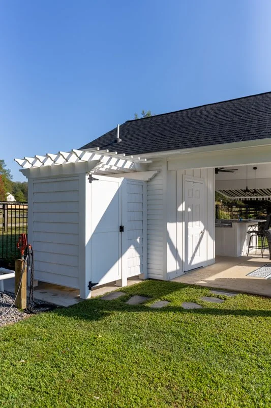 White shed with double doors and a small roof overhang, adjacent to a house with a porch, on a green lawn under a clear blue sky.