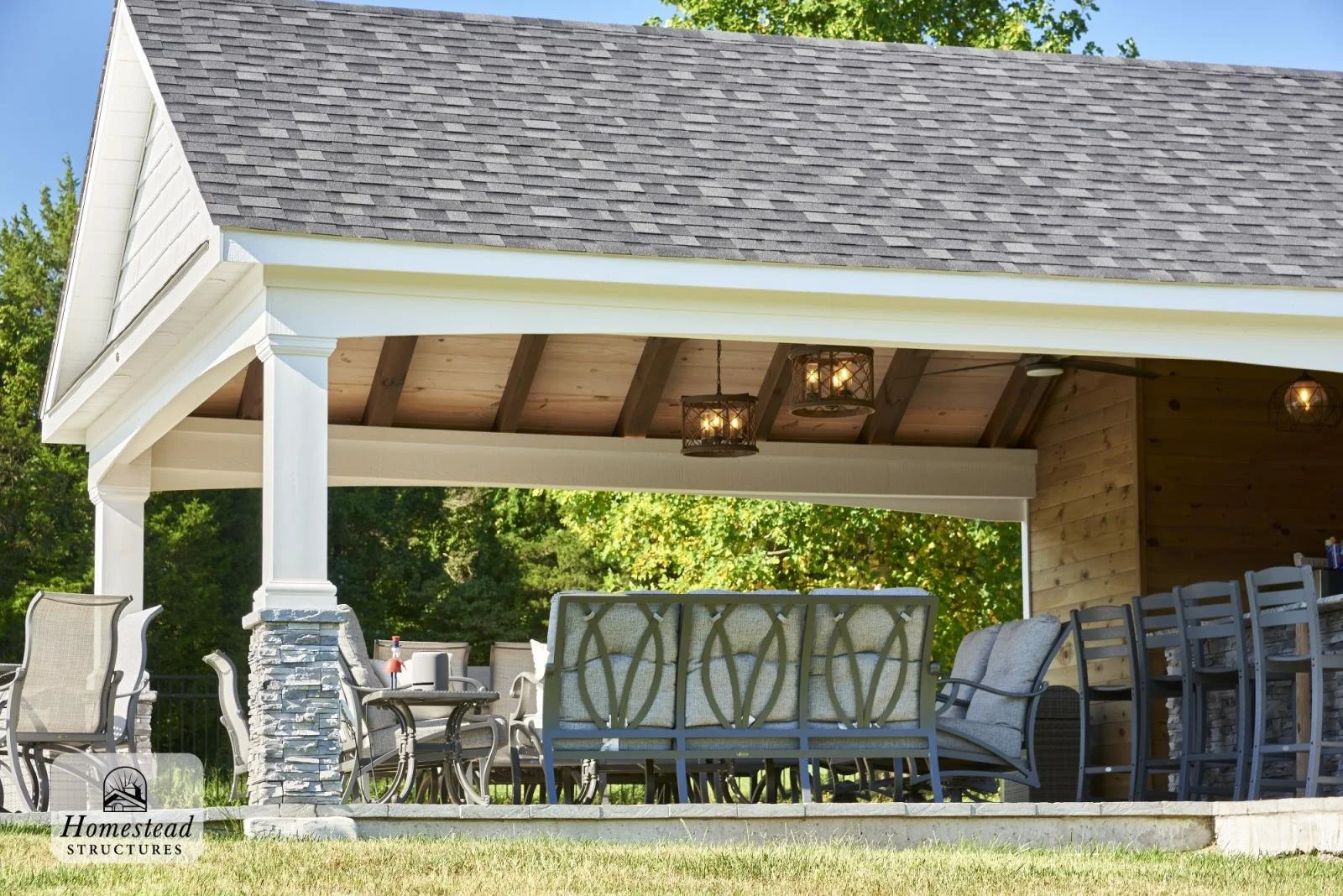 A covered outdoor patio with seating and dining area, featuring a vaulted ceiling with hanging light fixtures, surrounded by trees and greenery.
