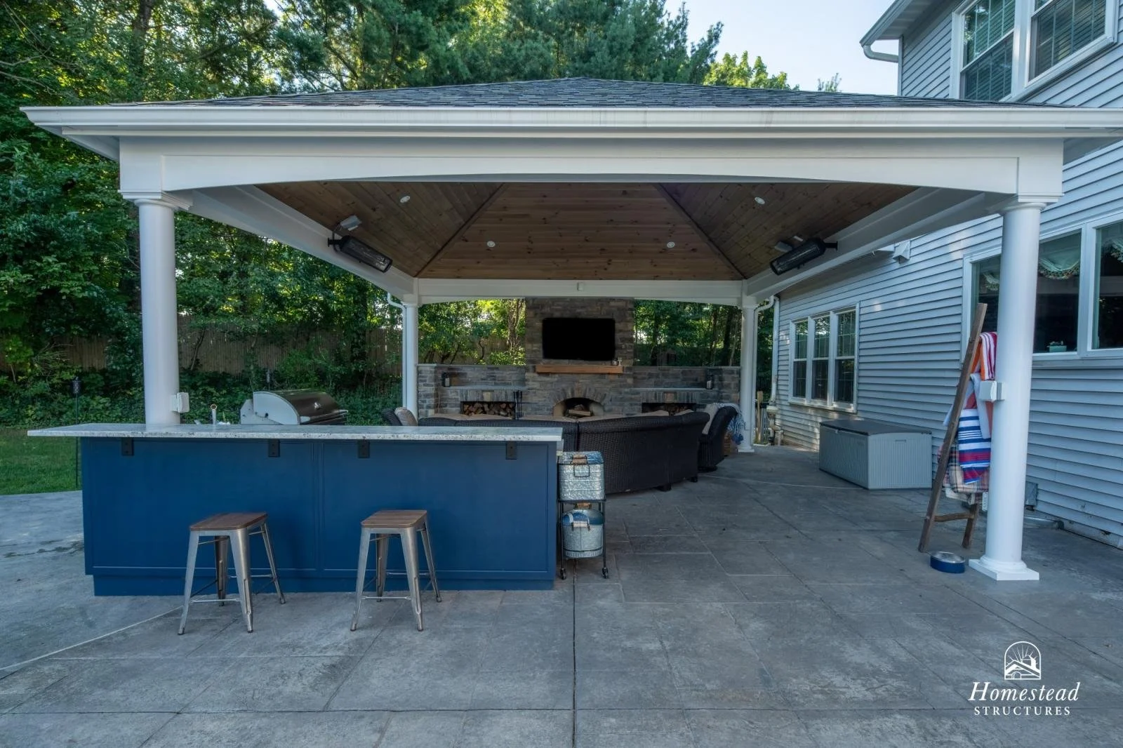 Outdoor covered living area with a kitchen island, seating, a fireplace, a TV, and string lights, surrounded by trees.