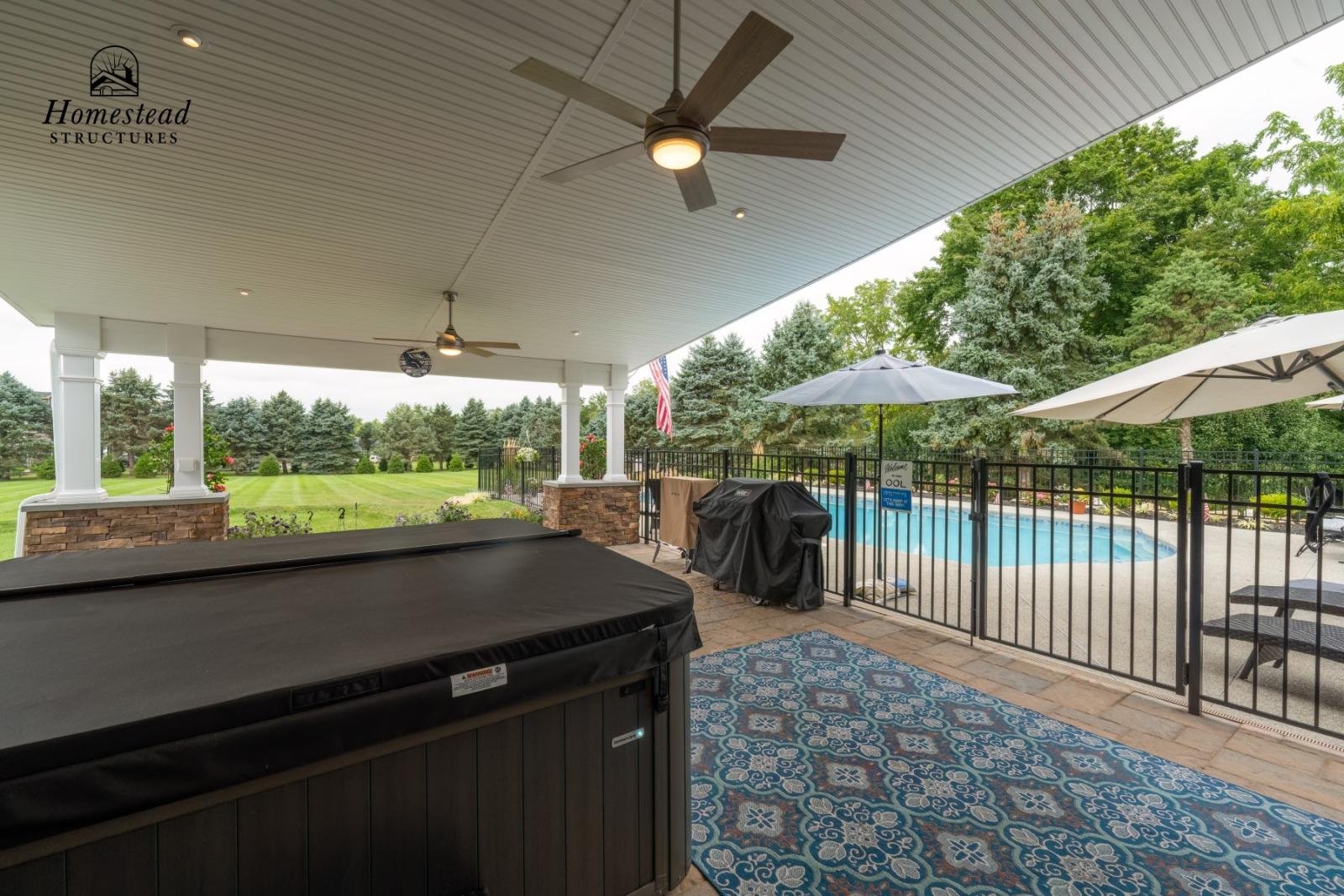 Covered patio area with hot tub, ceiling fans, and outdoor furniture overlooking a backyard with a swimming pool, umbrellas, and lush green trees.