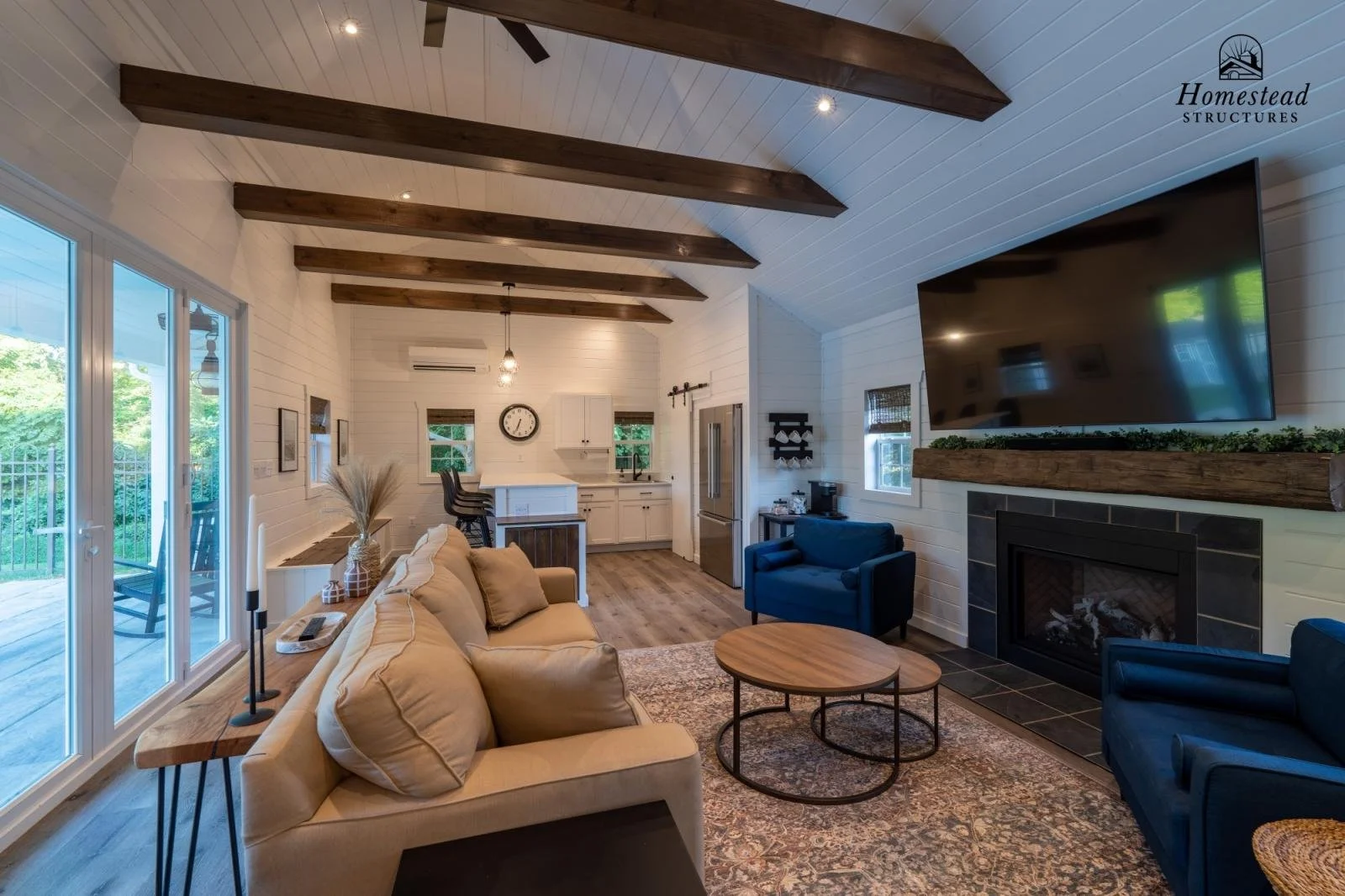 Living room with a beige sofa, blue armchairs, round wooden coffee tables, a fireplace with a large TV above it, a kitchen with white cabinets, a black clock, and wooden beams on the ceiling. Large windows and sliding glass door let in natural light.
