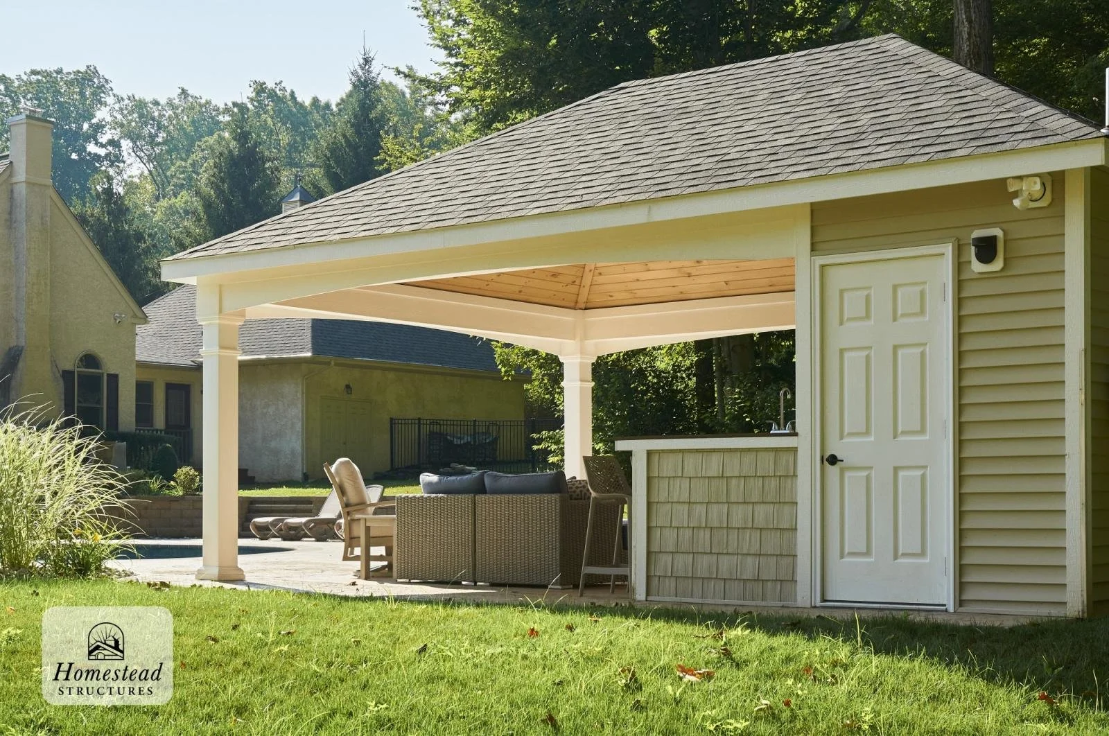 An outdoor backyard with a covered patio area featuring outdoor furniture, a small counter or bar, and a grassy lawn. There are neighboring houses and trees in the background.