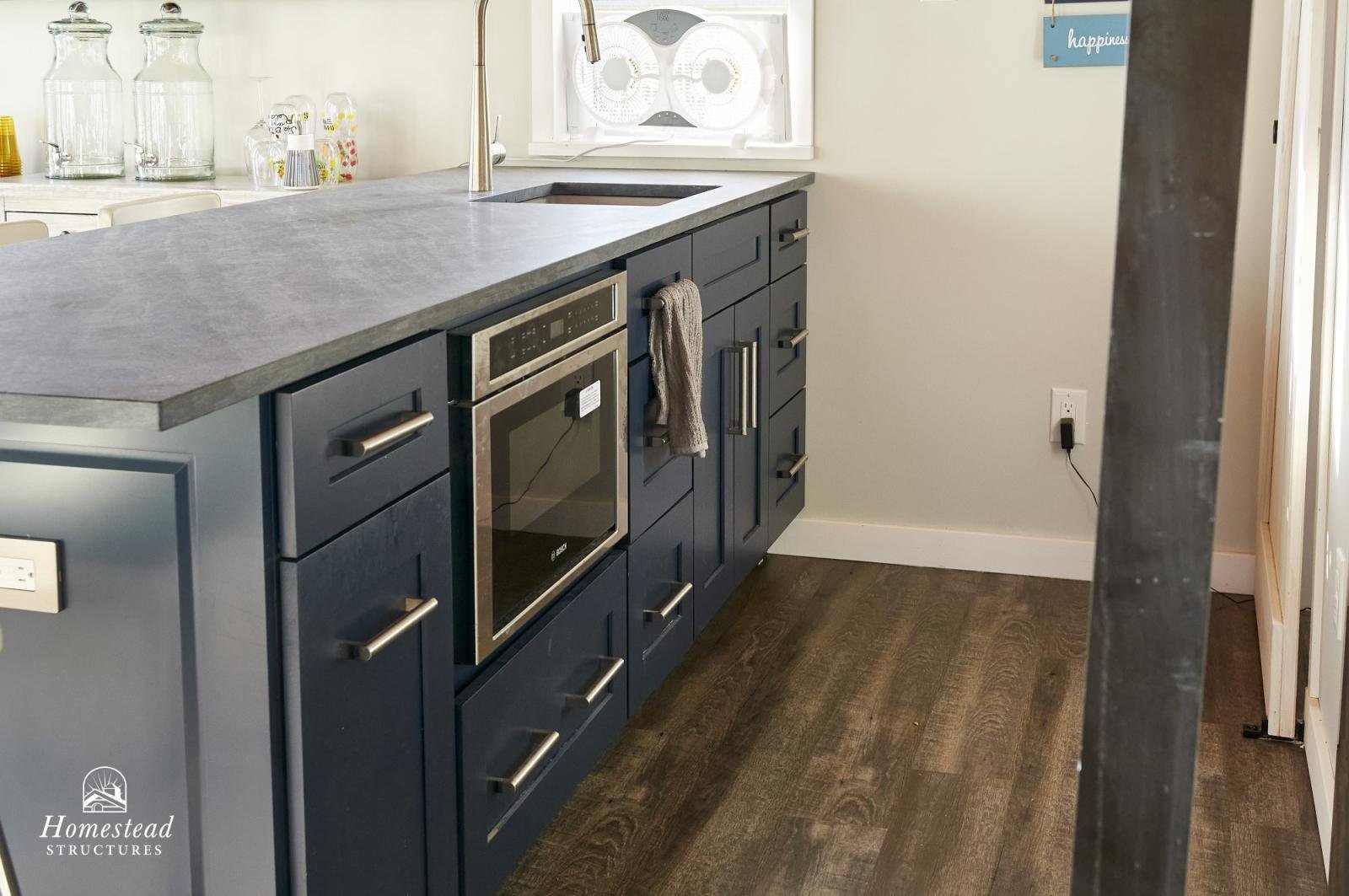 Photo of a modern kitchen with dark blue cabinets, a gray countertop, a built-in oven, a faucet, and a window with a fan. There is a towel hanging on a cabinet handle, and a power outlet on the wall.