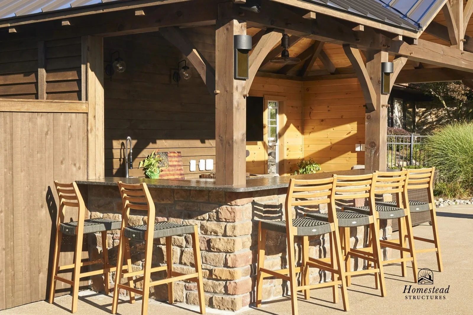 Outdoor bar area with a countertop, wooden bar stools, and a wooden structure. The background includes a wooden wall, some greenery, and exterior lighting.