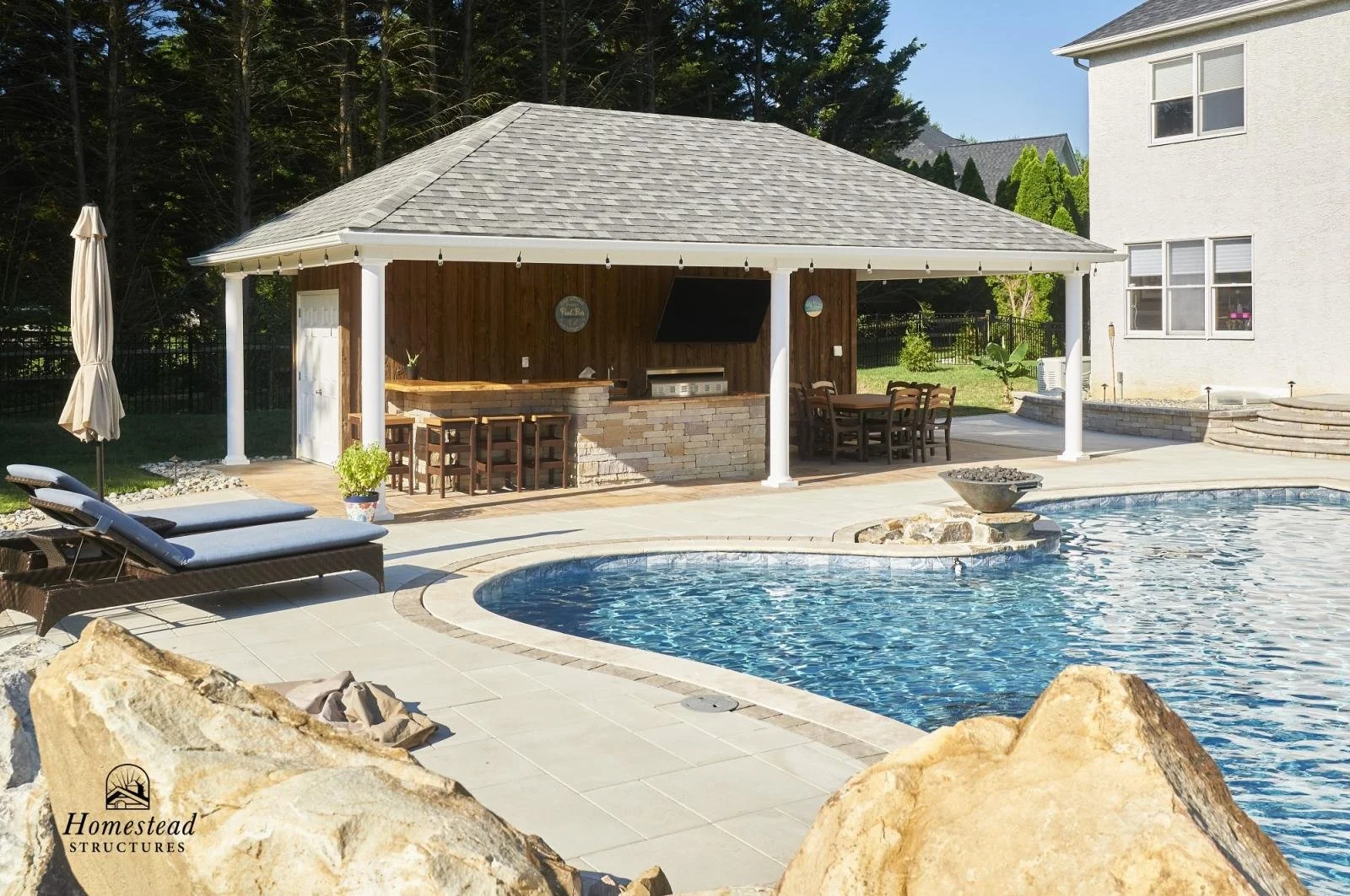 An outdoor pool area with a pool, lounge chairs, and a covered outdoor kitchen and dining area with a bar, grill, and table, surrounded by rocks and trees.