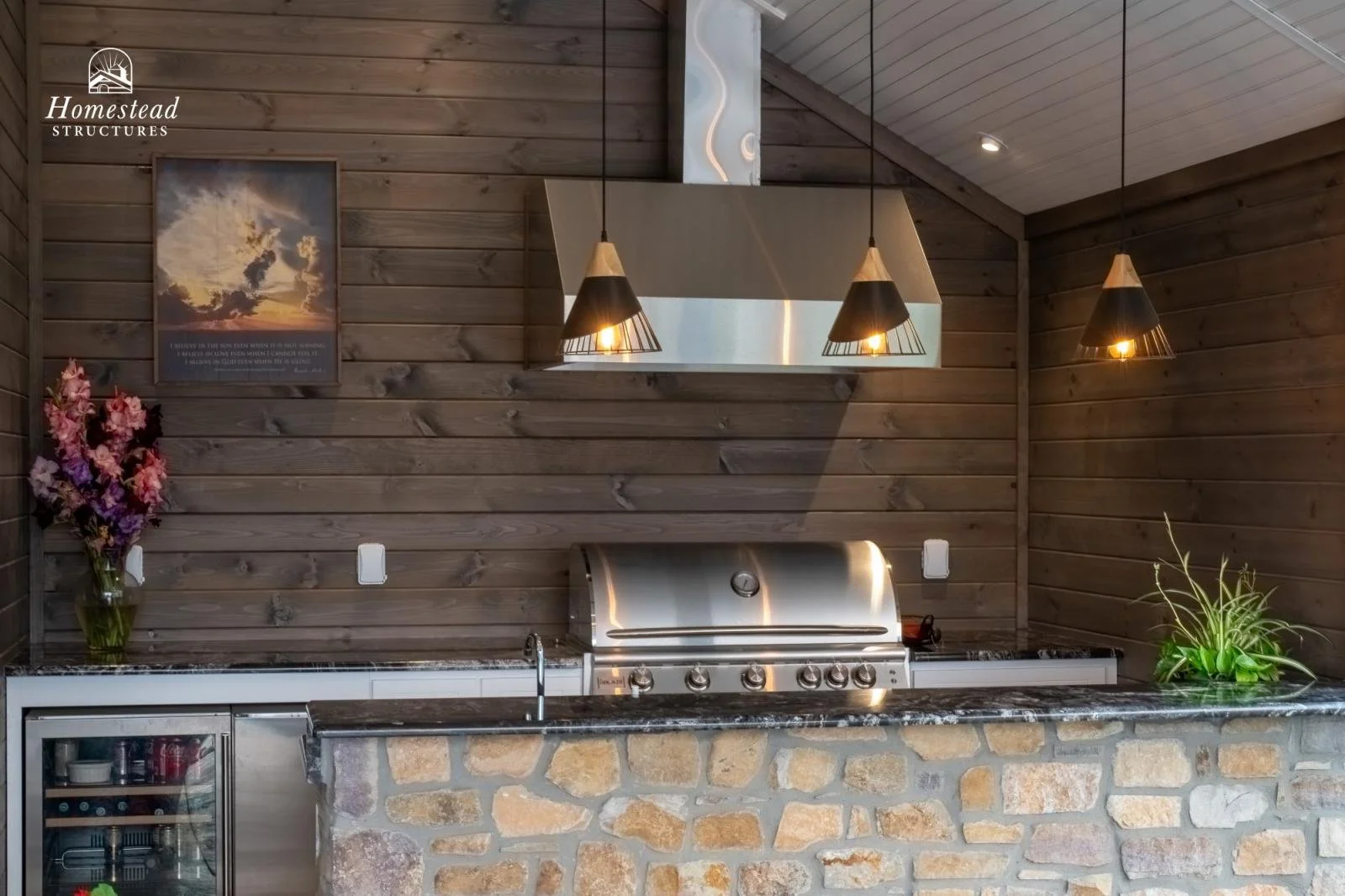 Outdoor kitchen with stone counter, stainless steel grill, wood-paneled walls, pendant lights, framed art, flower vase, and green plant.