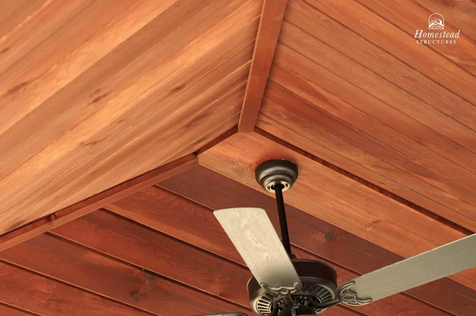 Close-up of a ceiling fan with black body and silver blades mounted on a wooden ceiling with reddish-brown planks.