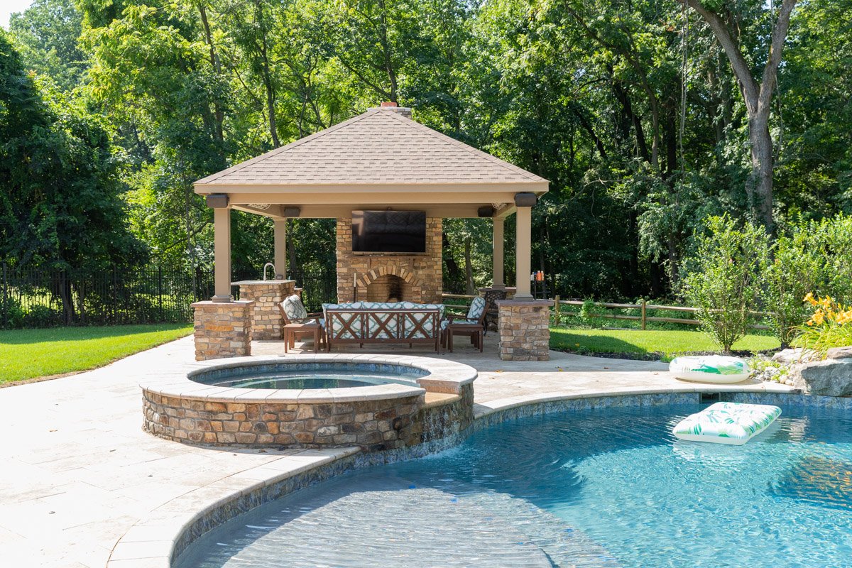 Backyard with a swimming pool, hot tub, and a shaded outdoor living space with a brick fireplace and a mounted TV, surrounded by lush green trees.