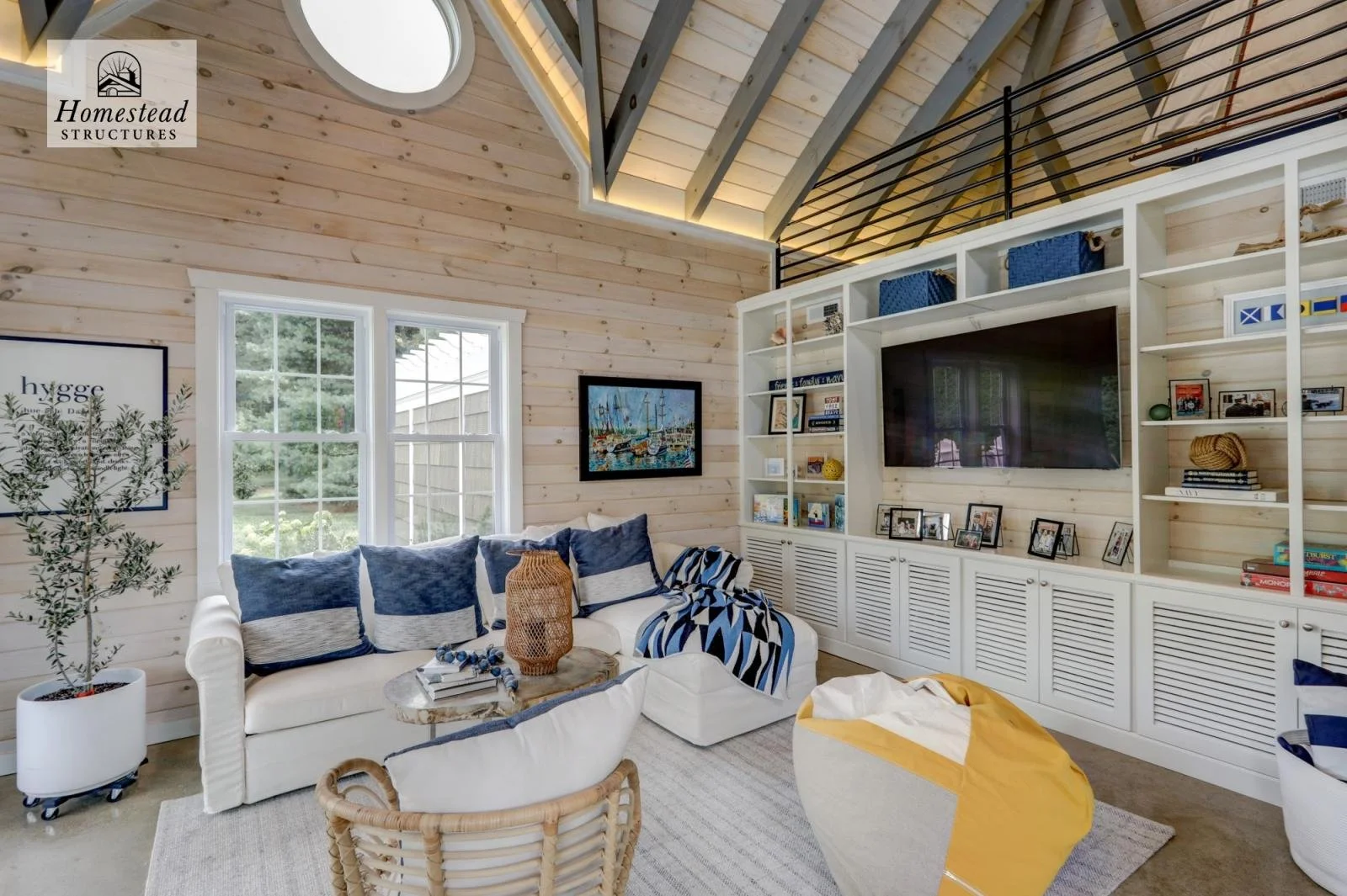 Living room with natural wood walls, a white sofa with blue and gray pillows, a wicker basket, a beige and yellow bean bag, a round coffee table, a large built-in white shelf with a TV, and black railing on the upper level. Two windows, a framed artw