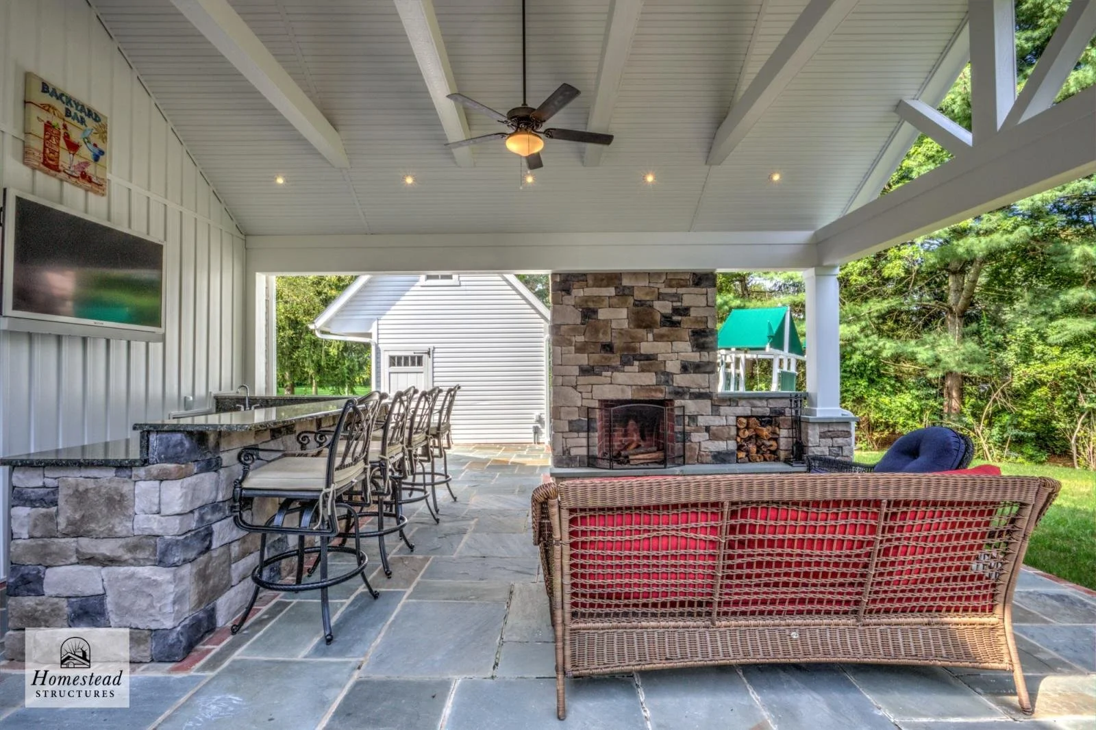 Covered outdoor patio with a stone fireplace, a wicker sofa with red cushions, a bar counter with stone facade and bar stools, a ceiling fan, and a view of trees and neighboring houses.