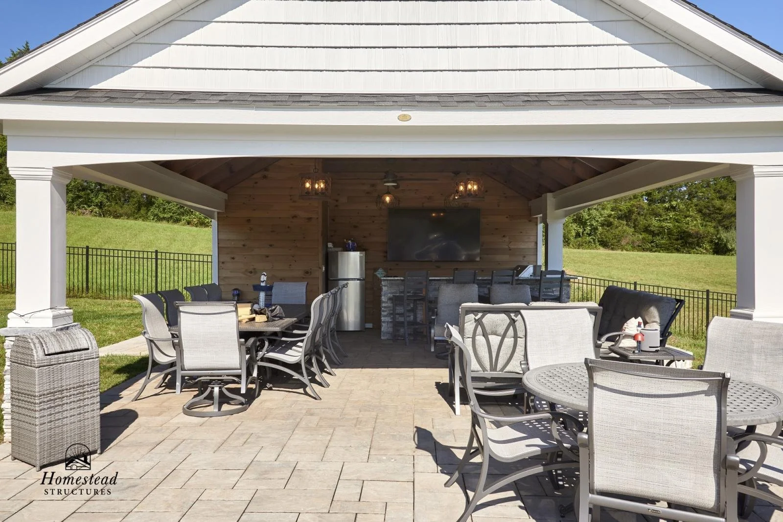 Outdoor covered patio with dining tables, chairs, and a bar area with a flat-screen TV, surrounded by a grassy yard and a black metal fence.