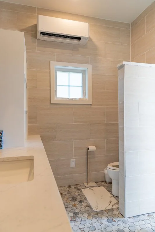 A modern bathroom with beige tiled walls, a small window, a wall-mounted air conditioner, a toilet with a white lid, a toilet paper holder, and a marble bath mat on a hexagonal tiled floor.
